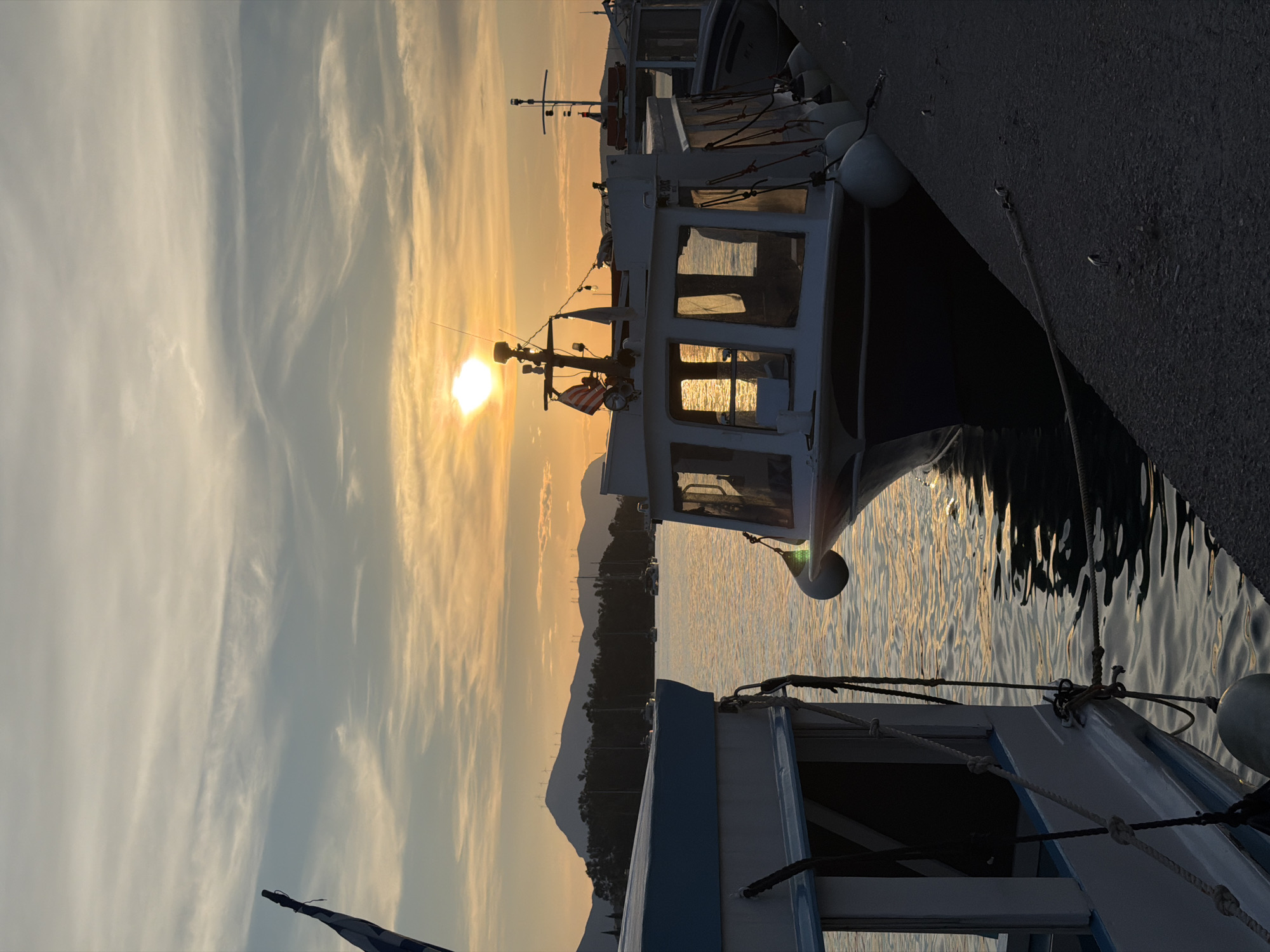The image captures a serene sunset scene at a harbor, likely in Poros, Greece. The sun, a bright golden orb, is positioned low on the horizon, casting a warm, golden glow across the sky and water. Wispy, elongated clouds stretch across the sky, transitioning from bright gold near the sun to soft gray-blue further away.
In the mid-ground, a white fishing or small passenger boat is prominently docked, its cabin windows reflecting the brilliant sunset light. On its mast, a small flag, possibly an American flag with red and white stripes, is visible. The boat is moored to a pier on the right, with large white buoys hanging from its side.
The calm sea reflects the golden hues of the sky, creating shimmering patterns on the water's surface. In the foreground, part of another boat, predominantly white and blue, is visible, with ropes and mooring lines neatly secured. In the distance, a dark silhouette of mountains or landmasses stretches across the horizon, providing a dramatic backdrop to the vibrant sky. The overall atmosphere is one of tranquility and natural beauty at dusk.