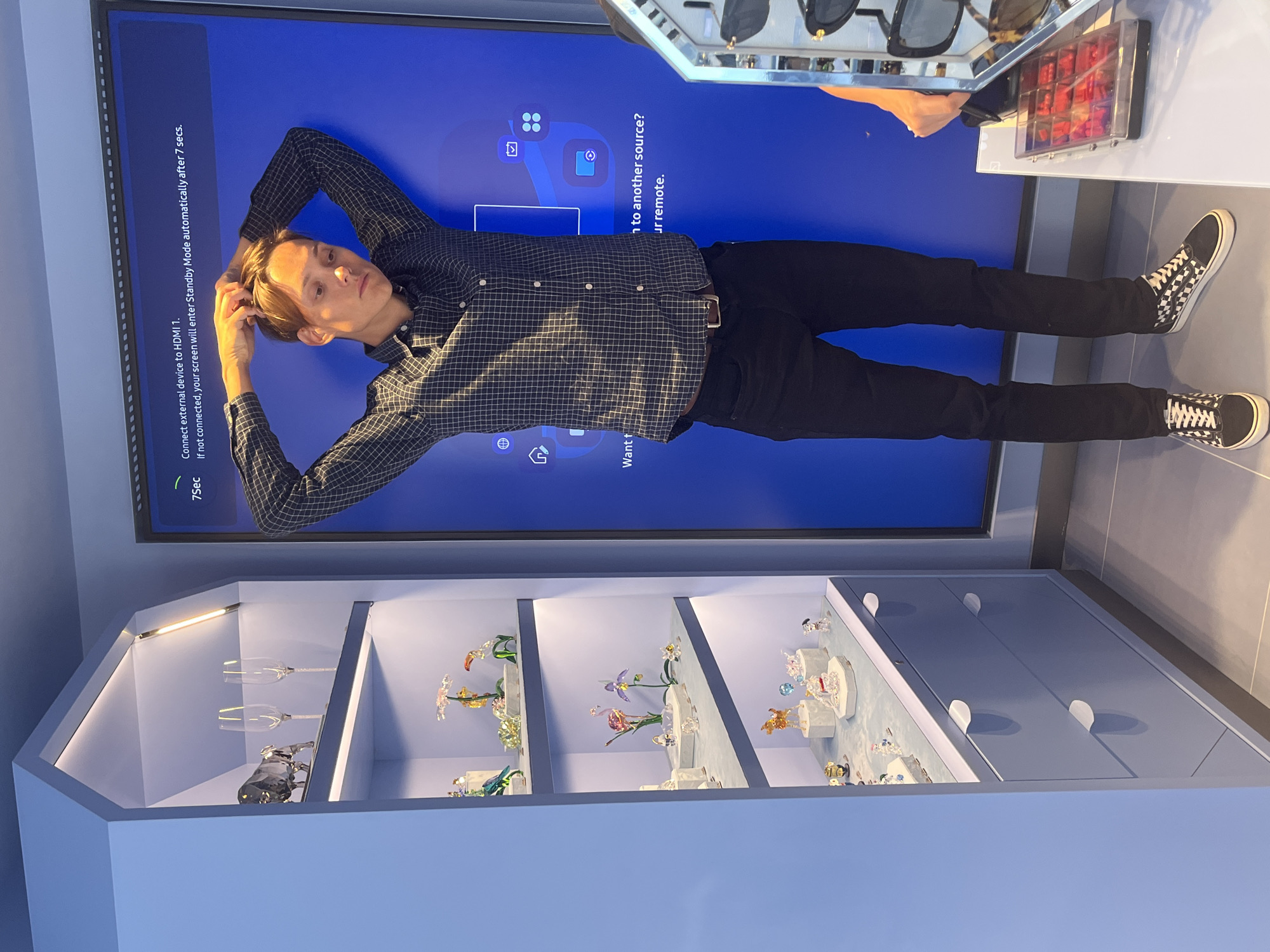 The image shows a young person lying down on the floor in front of a large, dark blue screen. The screen displays text and icons related to device connection and input sources. The person is wearing a checkered shirt, black pants, and black and white checkered Vans. Their arms are behind their head, and they are looking up towards the screen. The background shows a display cabinet filled with various glass figurines and two wine glasses. The lighting appears to be artificial, suggesting an indoor setting. The overall mood is relaxed and casual. The visible text on the screen reads: "Connect external device to HDMI 1." and "If not connected, your screen will enter Standby Mode automatically after 7 secs." Another section of text on the screen says "to another source? ur remote." There is also a visible timer displaying "7Sec". The setting appears to be a retail store or a showroom, possibly in Budva, Montenegro, given the location context. The time of day cannot be determined from the image.