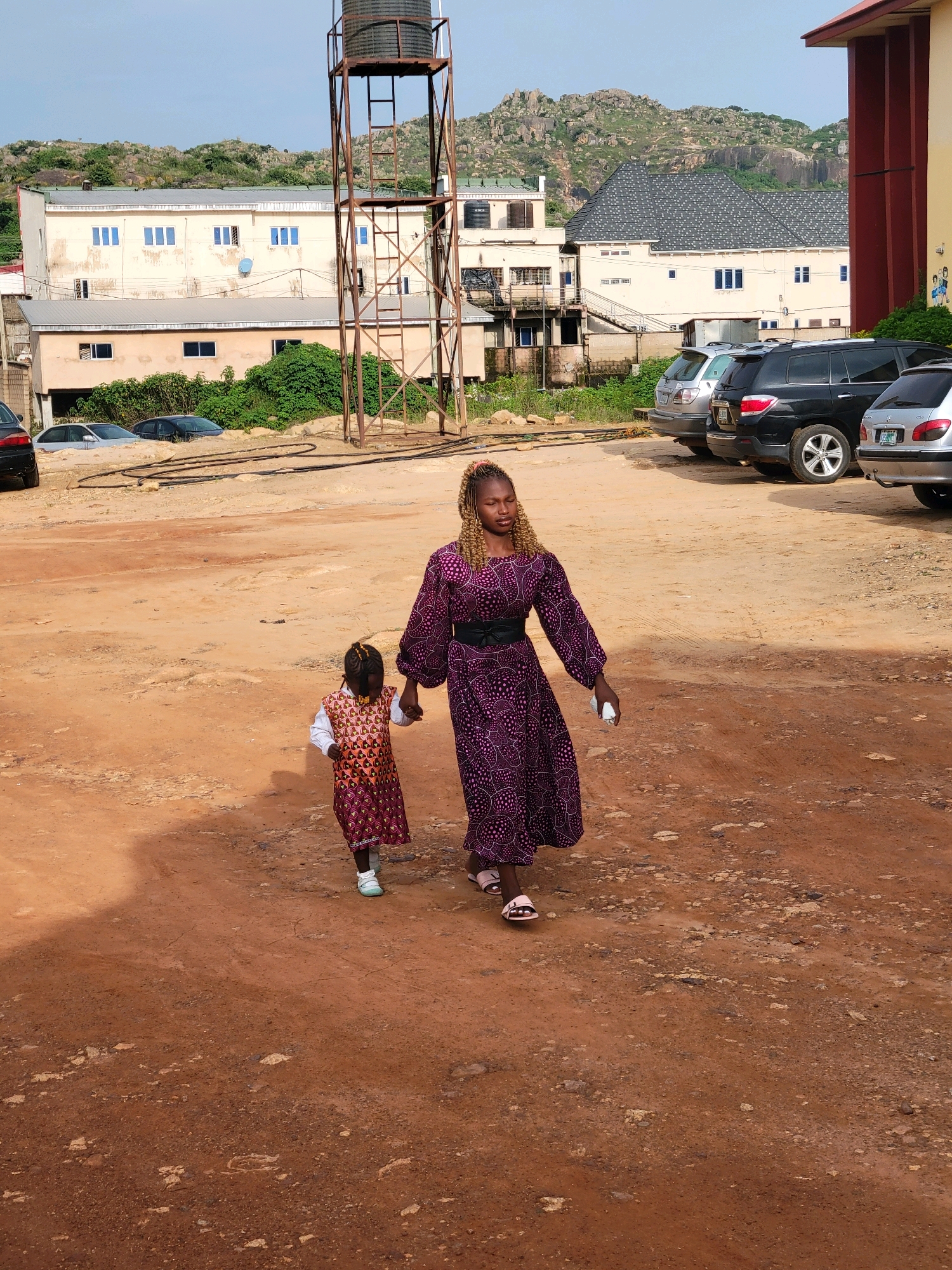 In Gura Top, Nigeria, a woman in a purple patterned dress walks hand-in-hand with a young child on a dirt path. The woman has blonde braided hair and is looking down, while the child, wearing a patterned dress over a white shirt, walks beside her. In the background, a water tower stands tall next to several multi-story buildings with light-colored facades and blue-framed windows. The area appears to be a parking lot with several parked cars, including a silver sedan, a black SUV, and another silver vehicle. Rocky hills and lush green vegetation are visible in the distance under a partly cloudy sky.