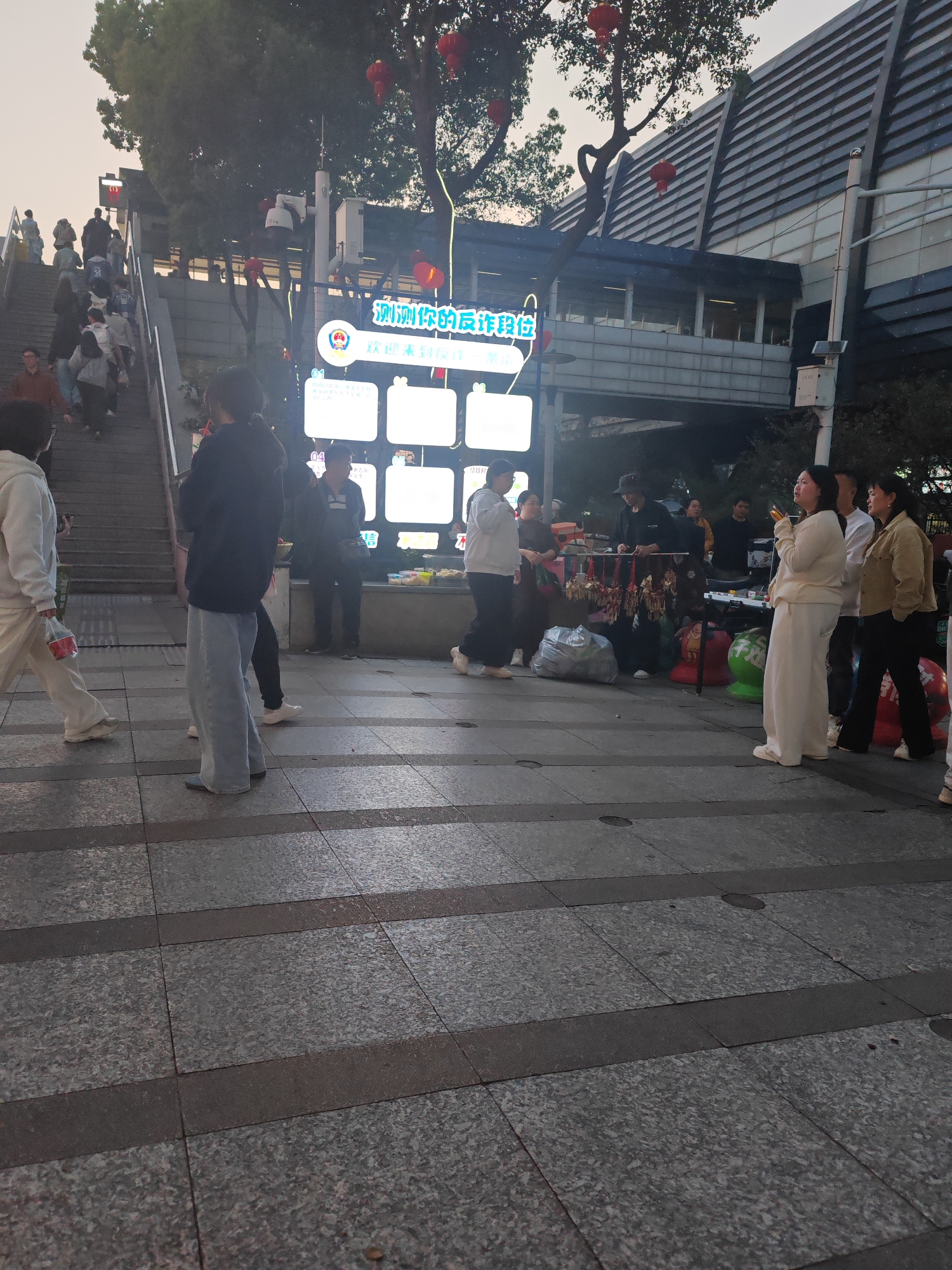 The image depicts a street scene in Jiangning District, China, with several people present. On the left, individuals are ascending a set of stairs. In the center, people are gathered around a display of illuminated screens with Chinese characters. Additional people are interacting with a stall featuring various items for sale, including decorations. The architecture in the background includes a modern building with a distinctive facade. The ground is paved with grey tiles. Red lanterns are visible hanging from trees.
