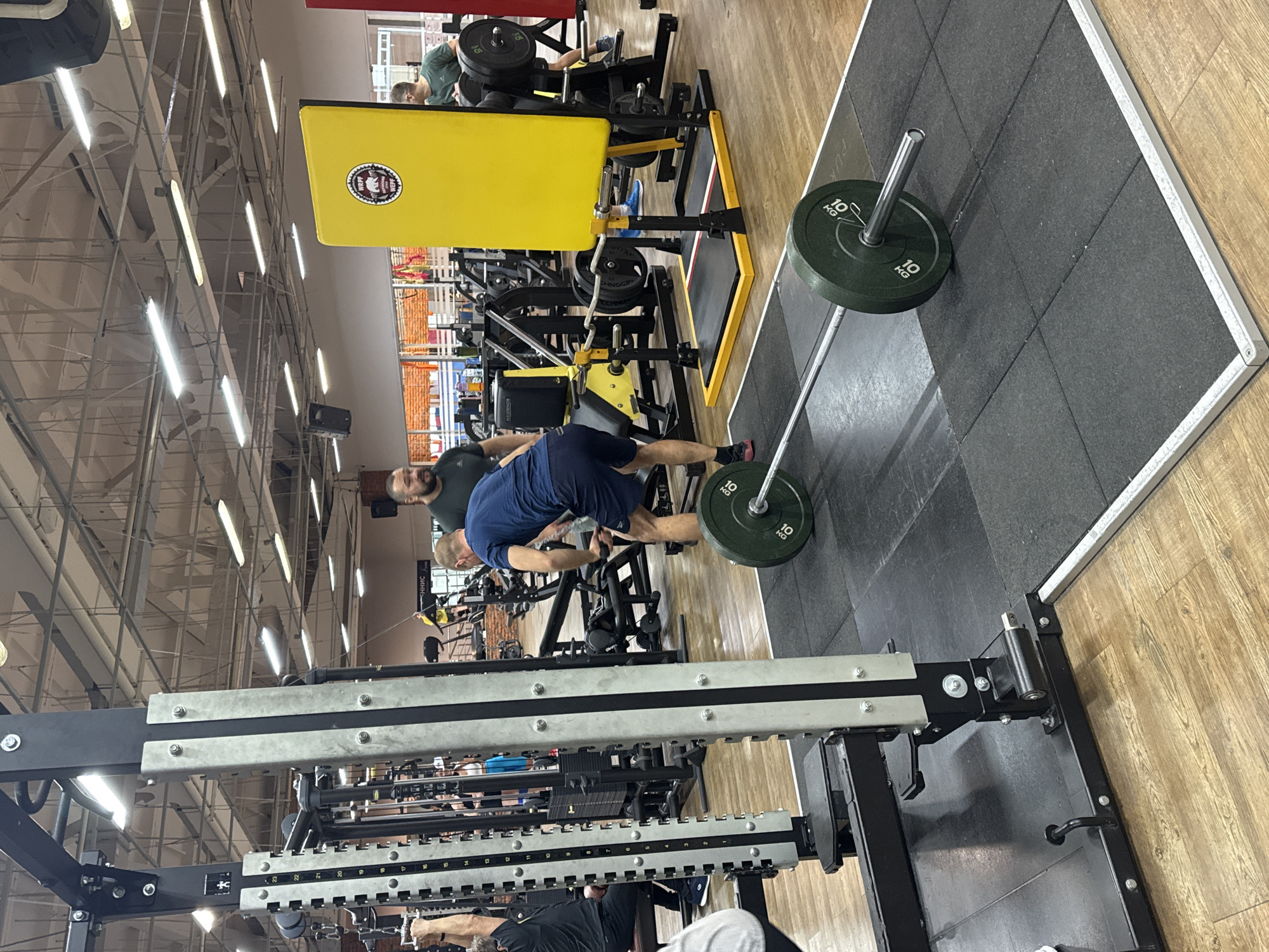 The image depicts the interior of a gymnasium in Chaikovsky, Russia. The gym is filled with various weightlifting machines and equipment. A man wearing a blue shirt is bending over to lift a barbell loaded with 10 kg weight plates. Another man with a beard is observing him. Other gym-goers are present, utilizing different machines in the background. The gym has a wooden floor with sections covered by black rubber mats. Overhead, the ceiling is fitted with numerous light fixtures.