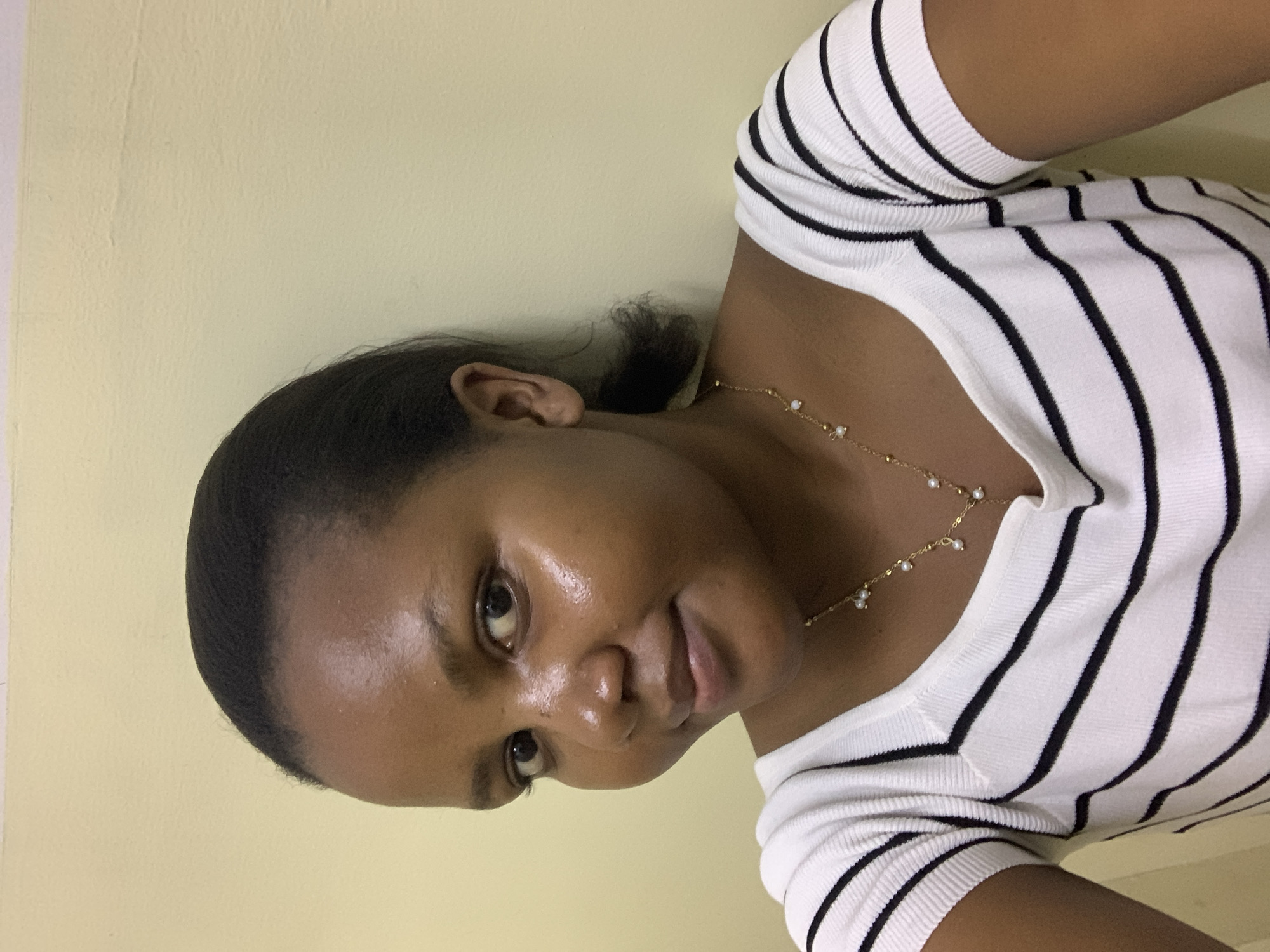 A young woman is pictured from a low angle, likely from a selfie, against a pale yellow wall. She has dark hair pulled back and is wearing a white, short-sleeved top with black stripes, and a delicate gold necklace with small pearl-like embellishments. The lighting seems to be artificial, and there are no indications to indicate that the image was taken in the city of Dar es-Salaam, Tanzania.