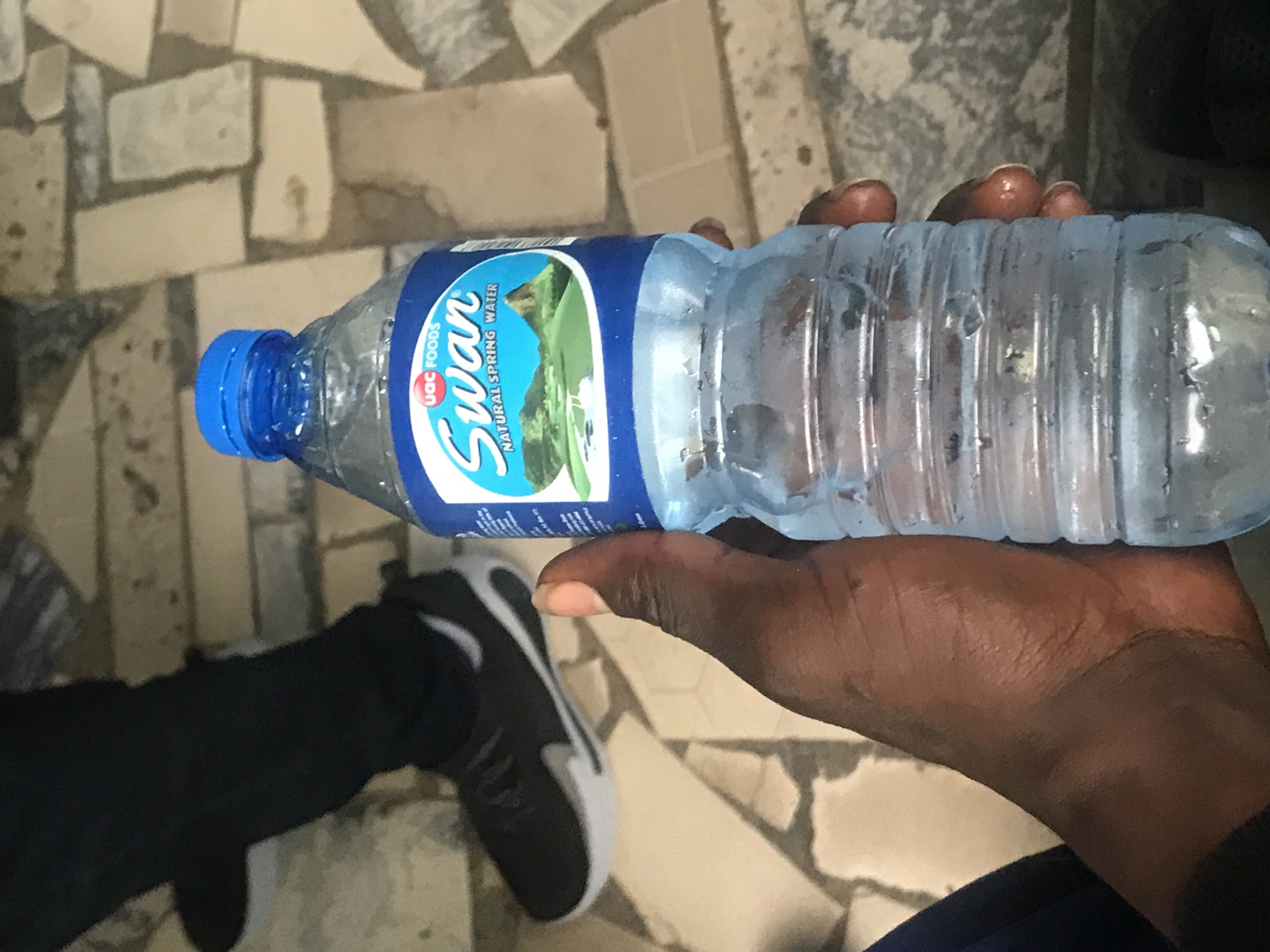 The image shows a person's hand holding a bottle of Swan Natural Spring Water. The bottle has a blue cap and a label featuring a landscape with mountains and a body of water. The hand is dark-skinned. In the background, a person wearing black pants and black and white sneakers is visible. The ground appears to be paved with irregularly shaped tiles. The scene suggests an outdoor setting, possibly a public area or walkway in Jos, Nigeria, given the context provided. The lighting indicates daytime.
