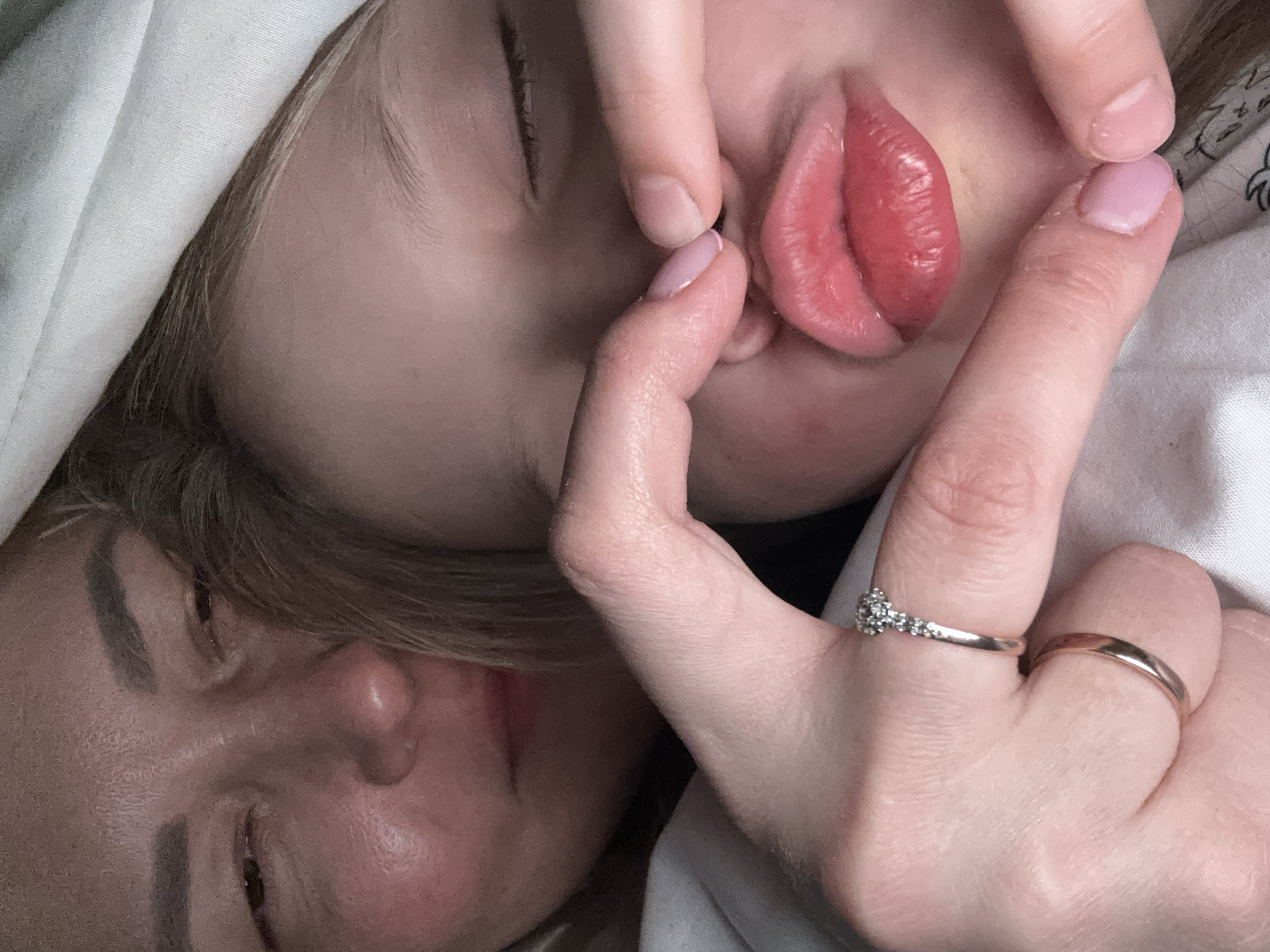 A close-up, intimate image shows two faces in close proximity. The upper face, seemingly that of a child, is partially obscured by fingers manipulating their lips, possibly stretching or pulling them. The child's eyes are closed. The hand manipulating the child's lips has a silver ring with small stones and a plain gold band on the ring finger. The lower face, partially visible, appears to be that of an adult, also with closed eyes. The background is blurred and unclear, with white fabric or bedding visible. The location in the city of Izhevsk, Russia, cannot be determined from the image.