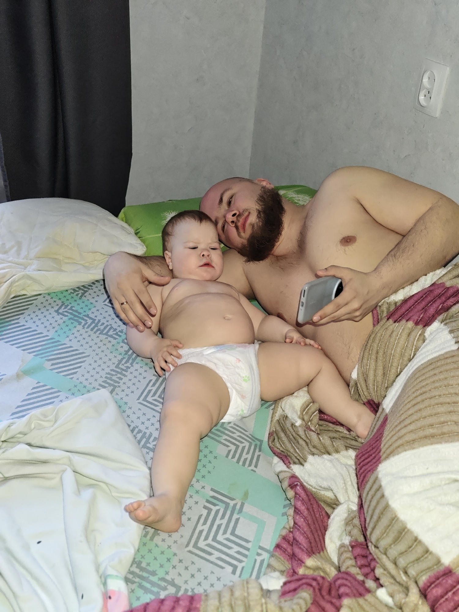 A man and a baby are lying in a bed. The man is shirtless and has a beard, and he is holding a smartphone. The baby is wearing a diaper. The bed has a patterned sheet and a blanket. In the background, there is a wall with an electrical outlet. The location of the photo is in the city of Chaikovsky, Russia.