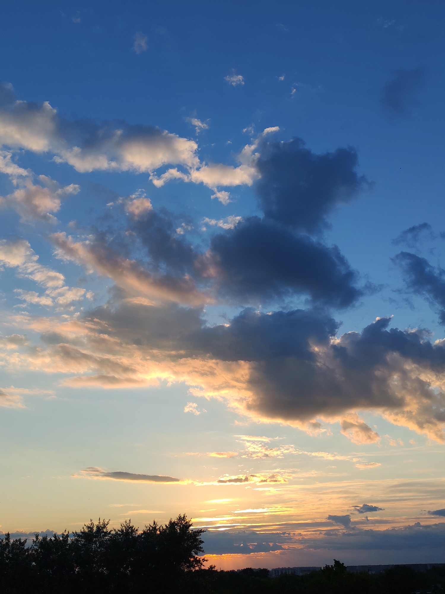 The media file displays a vibrant sky scene, captured during either a sunrise or sunset in Podolsk, Russia. The dominant subject is the expansive sky, filled with a dynamic array of clouds.
The upper portion of the image features a deep blue sky, scattered with smaller, brighter white clouds. As the eye moves downwards, the sky transitions into softer blues, then warm hues of orange and yellow near the horizon, indicative of the golden hour. The clouds in the middle and lower sections are illuminated by the low-hanging sun, showing shades of glowing orange, pink, and yellow, particularly on their edges, contrasting with their darker, shadowed undersides and larger grey formations. Some clouds appear wispy, while others are dense and volumetric.
The sun is visible as a brilliant glow just above the horizon, partially obscured by clouds and distant elements, casting warm light across the lower clouds. A dark, silhouetted line of tree tops stretches across the bottom of the image, suggesting a foreground of foliage. Below and behind the trees, a faint, dark outline of distant buildings or a cityscape is visible, blending with the horizon.
The overall impression is one of natural beauty and tranquility, highlighting the dramatic interplay of light and clouds. There is no visible text in the image.