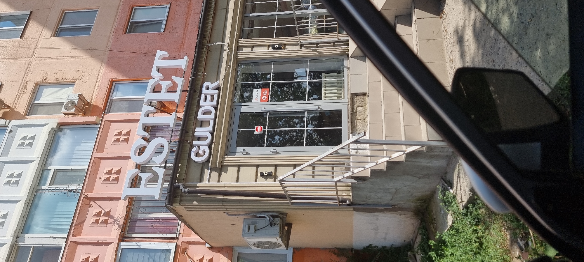 The image captures the storefront of "ESTET GULDER," a business located in Aktobe, Kazakhstan. The storefront is situated on the ground floor of a multi-story building with a salmon-pink facade. Prominent white, three-dimensional lettering spells out "ESTET" above and "GULDER" below. The entrance features double glass doors with a silver frame, flanked by windows also with silver frames. A metal railing with vertical bars leads up a short flight of tiled stairs to the entrance. An air conditioning unit is mounted on the wall to the left of the entrance. A red and white sticker, likely a QR code or informational sign, is affixed to the left door. The surrounding environment shows some greenery and cracked concrete at the base of the building, with a car's side mirror and part of its dark-colored frame visible in the foreground, obscuring the lower right portion of the scene. The sky is bright, suggesting daytime.