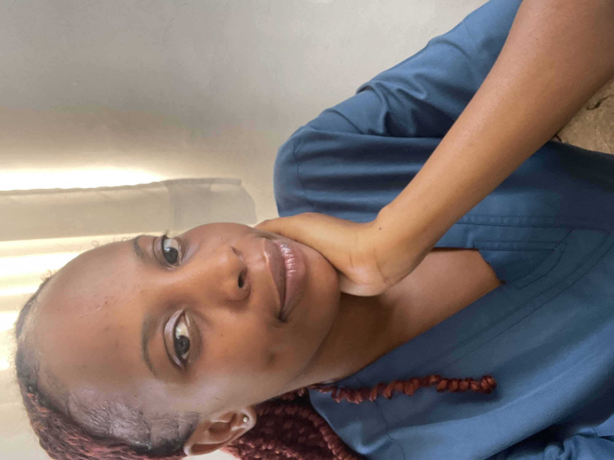 A young Black woman with reddish-brown braids is shown in a close-up, waist-up photograph. She is wearing a blue v-neck top. Her right hand is resting on her chin, and she is looking directly at the camera. Her left arm is visible, extended across the frame. The background consists of a textured wall and a window with sheer curtains, suggesting an indoor setting, likely a home or office, in Jos, Nigeria. The lighting appears to be natural daylight.