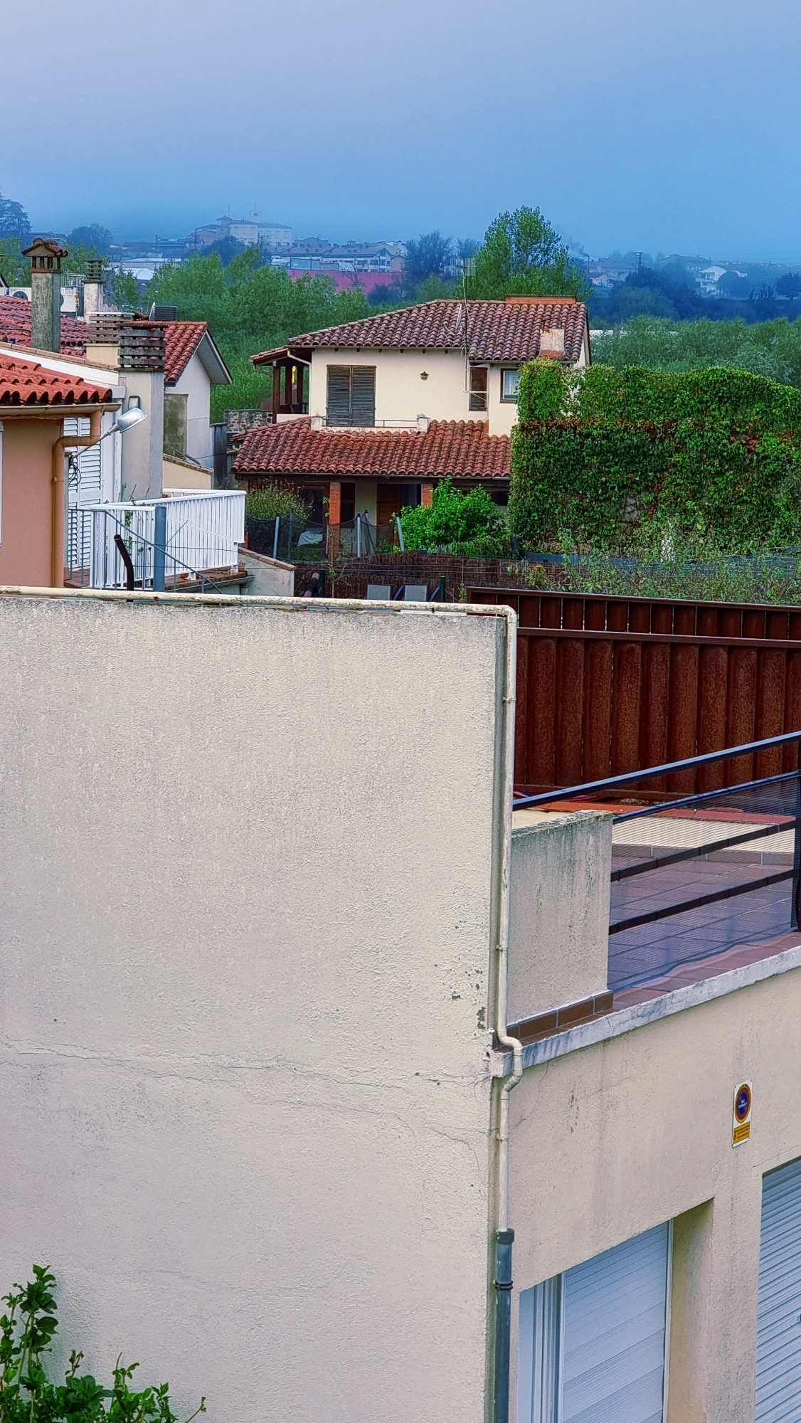 The image is an exterior shot of houses in the town of Sant Joan Les Fonts, Spain. The scene is dominated by terracotta tiled roofs and the muted, overcast sky, suggesting a cool or damp day. In the foreground, a light-colored, textured wall occupies the lower half of the frame. To the right of this wall, a balcony or terrace is visible, featuring a wooden deck and a dark railing. Behind this, a tall, vertically ribbed, reddish-brown metal fence runs horizontally across the mid-ground.
Further back, several houses are visible. One house, painted a pale yellow, has a prominent terracotta roof and dark-shuttered windows. To its left, other houses with terracotta roofs and chimneys are partially obscured by trees and vegetation. In the far distance, a hazy view of a larger town or city is visible, with buildings and faint outlines of structures blending into the mist-shrouded horizon. Green foliage, including a dense hedge on the right side of the frame and trees scattered throughout the scene, adds a natural element. The overall impression is a residential area in a somewhat subdued, perhaps rural, setting within Sant Joan Les Fonts.