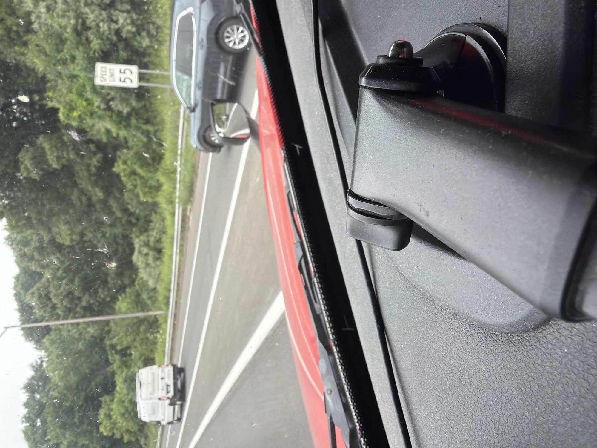 An eye-level, slightly high-angle, medium shot captures a view from inside a vehicle traveling on a multi-lane road. In the foreground, the dashboard of the vehicle is visible, with a black textured surface and a red dashboard accent. A black dashboard mount for a camera or electronic device is prominently featured in the lower right and center of the frame. Beyond the dashboard, the road ahead is visible with white lane markings.
In the mid-ground, a white van is traveling in the left lane, and further to the right, a dark-colored SUV is in the right lane. Both vehicles are moving away from the viewer. A speed limit sign indicating "55" is visible on the right side of the road, surrounded by lush green trees that line the highway. The sky appears overcast. The context of Danbury, United States of America, is not directly discernible from the visual elements of the image.