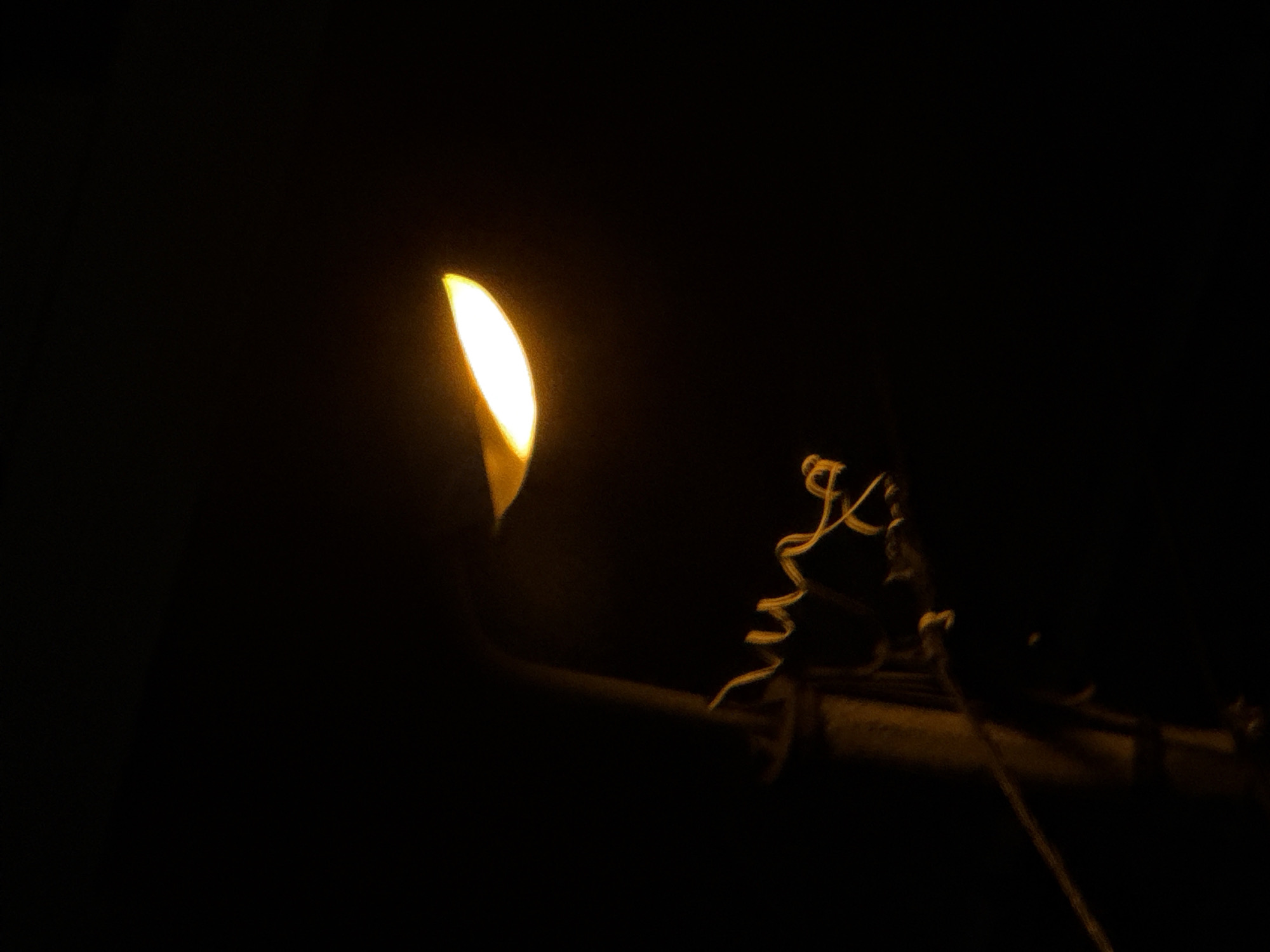 This image shows a close-up of a street lamp at night. The lamp is emitting a bright yellow light, casting a warm glow against the dark background. The lamp post is visible, made of a rough, textured material, likely concrete or stone. Wires are wrapped around the post, some coiled and loose. The scene appears to be outdoors, in an urban setting, possibly in Tbilisi, Georgia, given the prompt's context. There are no people or animals visible in the frame. The overall impression is one of a functional urban element in a dimly lit environment.