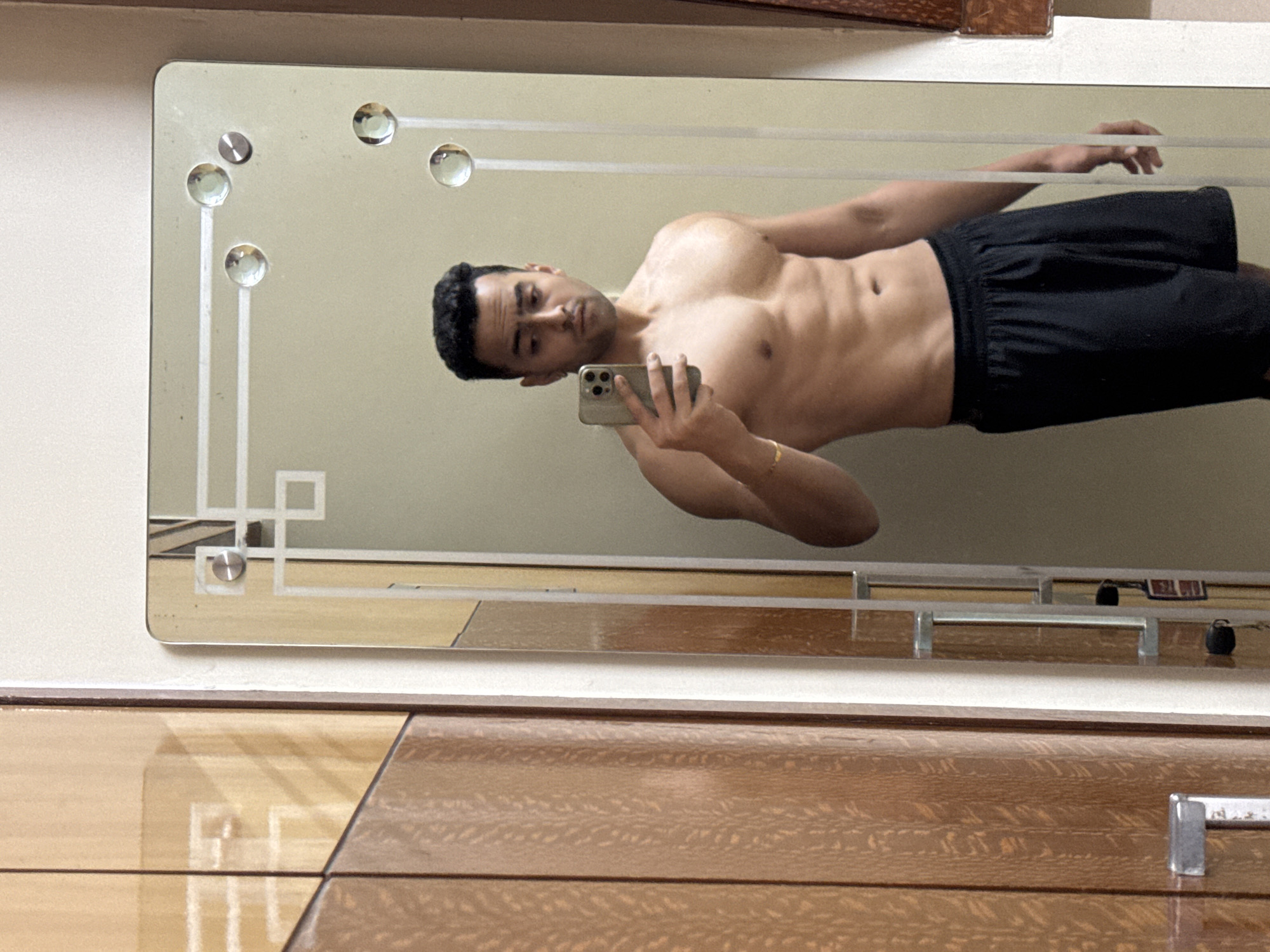 A man is depicted taking a mirror selfie indoors, appearing shirtless and wearing black shorts. He has dark hair, a light complexion, and a muscular build, with visible abdominal and chest muscles. He holds a smartphone with a multi-lens camera in his left hand, using it to capture his reflection. A gold-colored bracelet adorns his left wrist.
The man is reflected in a rectangular mirror, which is mounted on a plain, light-colored wall. The mirror features decorative etched patterns consisting of straight lines and geometric shapes, particularly in the lower left corner. Several circular, clear glass or acrylic embellishments are embedded in the mirror's surface, enhancing its decorative design. A metallic screw or fixture is visible near the top left corner of the mirror, securing it to the wall.
Below the mirror, a piece of wooden furniture, possibly a dresser or cabinet, is partially visible. It has a glossy finish with a distinct, light brown wood grain pattern. The polished surface of this furniture reflects parts of the room above it, including what seem to be small objects or elements from the mirrored area. The image is oriented sideways, with the man appearing horizontally. The setting suggests an indoor environment, possibly a bedroom or dressing area in Kolkata, India. No visible text is present in the image.
