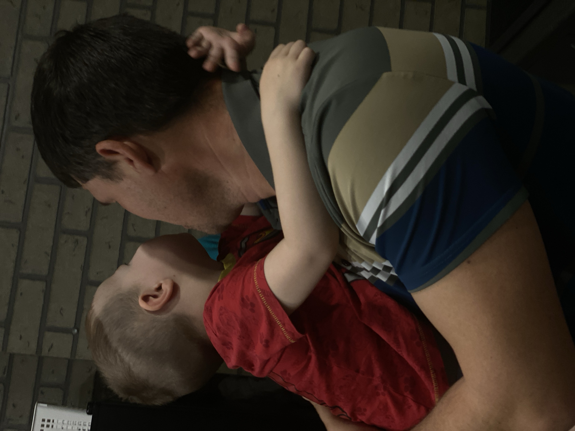 The image captures an adult male holding a young child in an embrace in Sochi, Russia. The adult male is seen from the back and right side, with short dark brown hair, wearing a multi-striped polo shirt featuring horizontal bands of dark grey, khaki, white, and blue. The young child, visible from the back and left side, has short, light brown hair with a shaved design on the side, and wears a red short-sleeved t-shirt with a faint pattern. The child's arms are wrapped around the adult's neck, and the child's head is tilted back and upward, positioned close to the adult's face. The background consists of a dark brick-patterned wall. To the far left, a partially visible white, perforated plastic container rests on a dark surface. The scene is indoors with subdued lighting.
