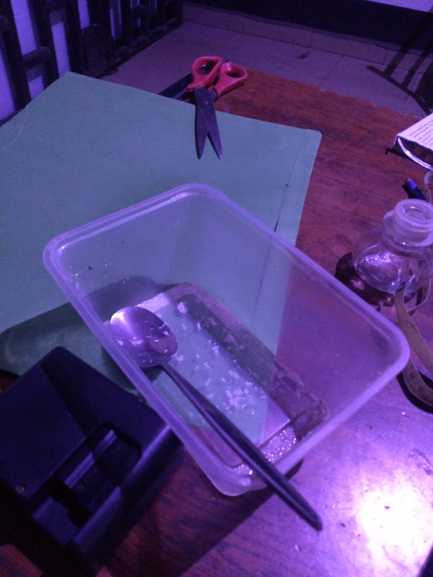 This image captures an indoor scene on a wooden table, illuminated by a pervasive purple-blue artificial light, set in Jos, Nigeria. No individuals are present. A large sheet of green material, consistent with craft foam or thick paper, occupies the left portion of the midground. A pair of red-handled scissors, with blades partially open, rests on the wooden surface above the green material, to the right.
In the center foreground, a clear, rectangular plastic container holds a silver spoon and exhibits white, granular residue on its bottom surface. To the lower left of this container, a black electronic device, possibly a power bank or a similar compact gadget, is positioned. On the right side of the table, behind the plastic container, a small clear glass bottle with a white stopper and clear liquid is visible. Portions of a yellow-brown measuring tape and a white package or paper with printed text are partially discernible near the bottle. The background displays a dark, possibly wooden or metal, latticework structure, further emphasized by the violet lighting. The arrangement of objects suggests a temporary workspace or a collection of craft-related items.