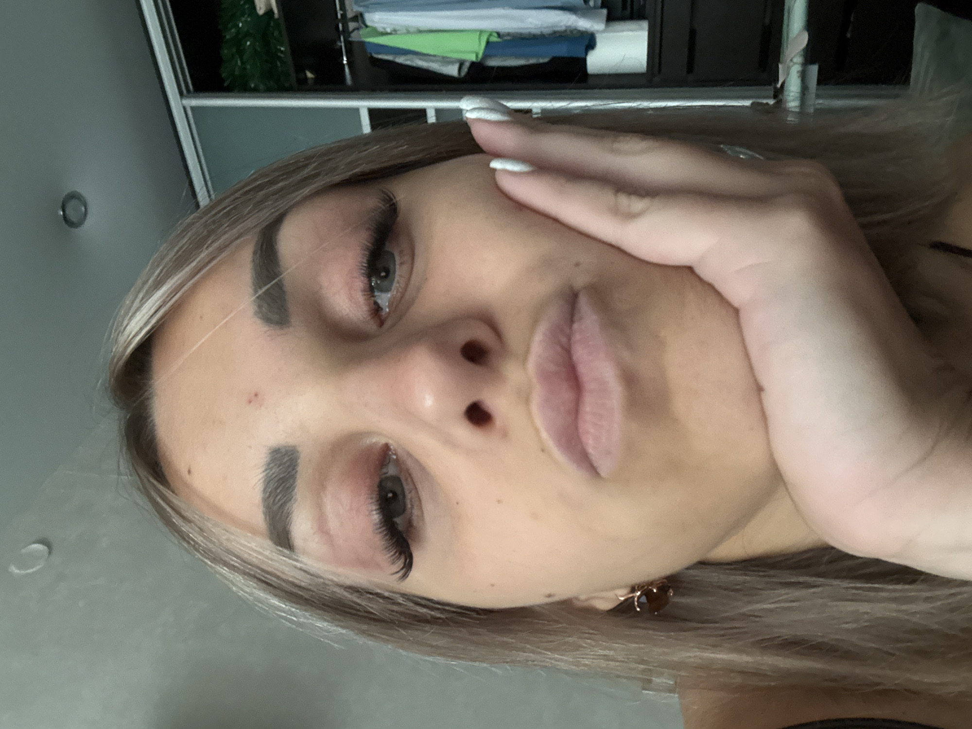 A young woman with blonde hair and dramatic false eyelashes is shown in a close-up selfie. She is resting her chin on her hand, and her mouth is pursed. A closet with clothing is visible in the background. The woman is wearing an earring. There is a red mark on her forehead. The image is a selfie taken indoors. There is no discernible indication of location in Yuzhno-Sakhalinsk, Russia.