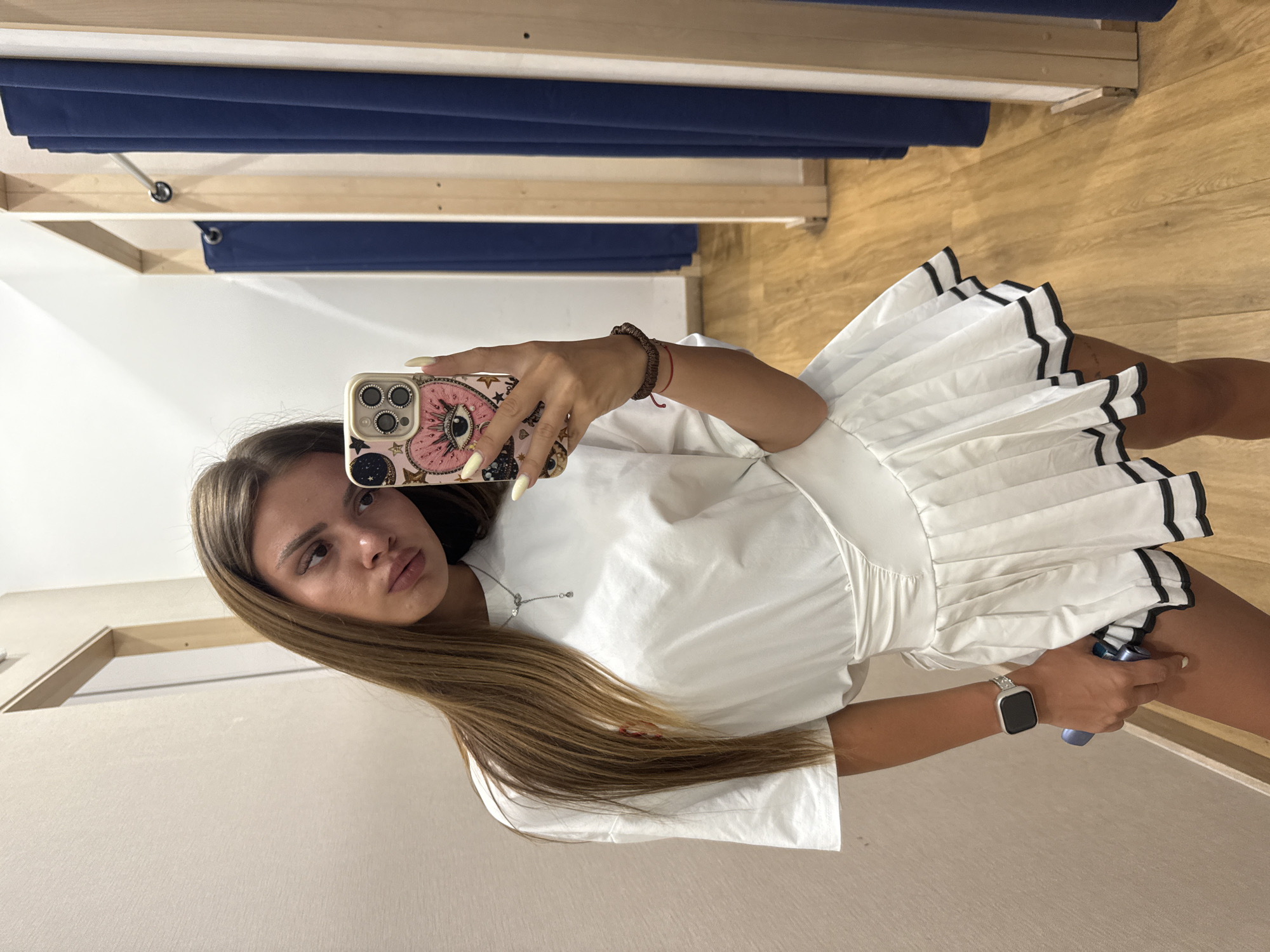 A young woman with long, brown hair is taking a selfie in what appears to be a fitting room. She is wearing a white t-shirt and a white pleated skirt with black trim. She is holding a smartphone with a decorated case in her right hand, and her left hand is resting on her hip. A smartwatch is visible on her left wrist. The fitting room has light-colored walls and a wooden floor. Blue curtains are visible in the background, suggesting the presence of multiple fitting stalls. The lighting is bright, likely from overhead fixtures.