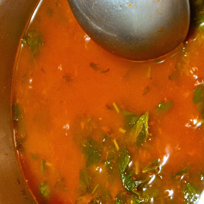 The image provides a close-up, top-down view of a vibrant reddish-orange liquid food in a pot, with a metallic ladle partially submerged within it. The scene captures an intimate moment of food preparation or serving.
The main subject is a thick, opaque liquid, likely a soup, stew, or curry, characterized by its rich, warm reddish-orange hue. This color suggests a tomato-based, paprika-rich, or chili-infused dish. Scattered generously throughout the liquid are numerous pieces of dark green, leafy ingredients, varying in size and shape. These appear to be herbs such as cilantro, parsley, or basil, or possibly finely chopped green vegetables, and some show signs of being cooked or wilted. Smaller, darker specks are also visible, likely ground spices or other finely minced ingredients. The surface of the liquid exhibits tiny bubbles and slight variations in texture, suggesting it is warm or recently cooked.
A large, reflective metallic ladle, likely made of stainless steel, occupies a significant portion of the upper right and central area of the frame. Its rounded bowl is partially submerged in the liquid, with light reflecting off its curved surface.
The edge of the cooking vessel is visible along the left and bottom perimeter of the image. It appears to be the inner wall of a pot, possibly made of metal or a ceramic-coated material, with a light brown or beige tone. Faint black markings are discernible on the inside wall of the pot, near the bottom left corner. Specifically, the text "1.5L" is visible, indicating a volume measurement on the pot's interior.
The lighting appears bright and somewhat diffused, illuminating the food and creating highlights on the ladle and the surface of the liquid. The overall impression is one of a hearty, flavorful dish being prepared or about to be served. No people are visible in the frame.