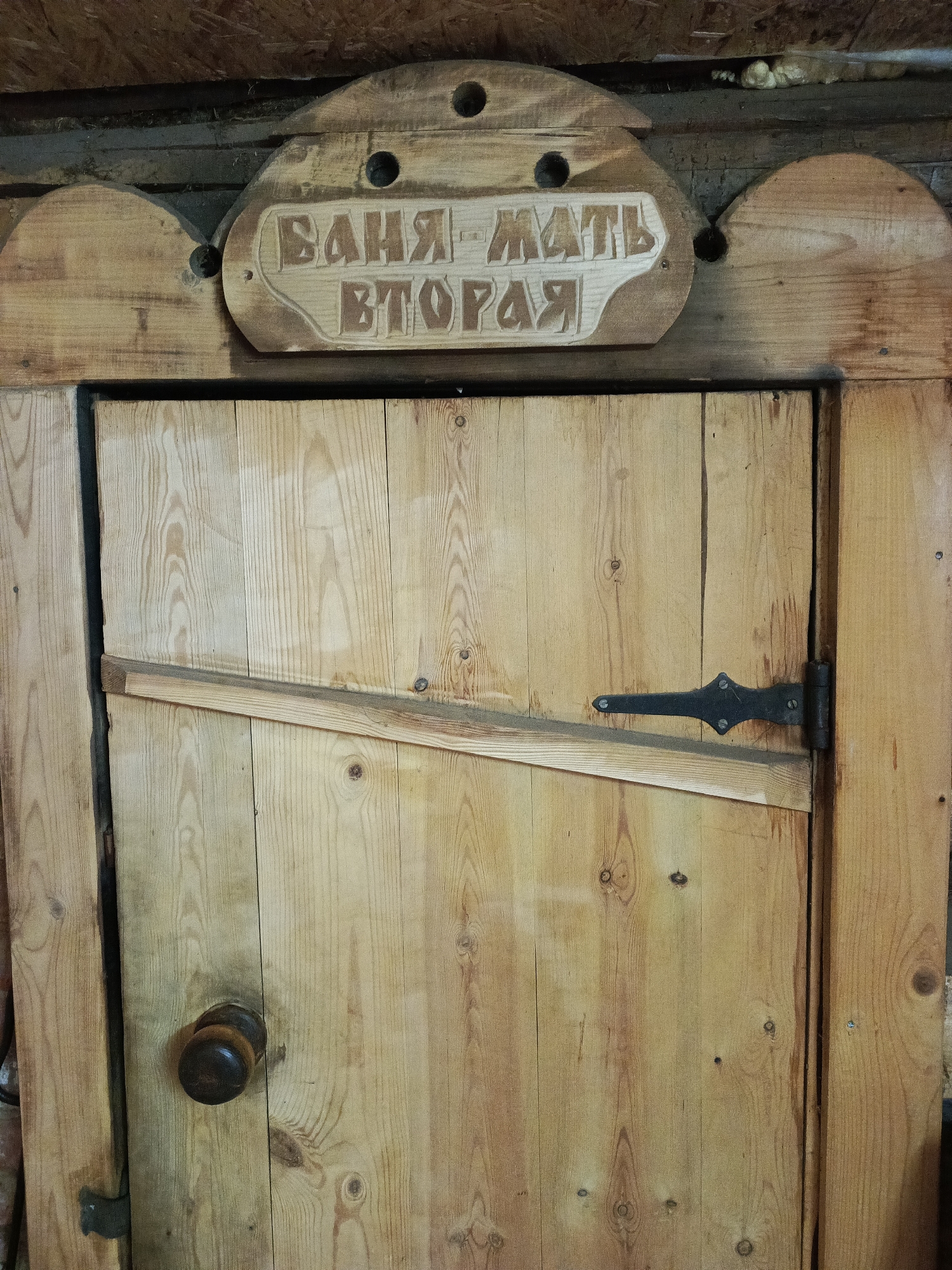 The image shows a wooden door in what appears to be a rustic or rural setting. A sign above the door, written in Cyrillic, reads "БАНЯ - МАТЬ ВТОРАЯ" - which translates to "Bath - Mother Second." The door itself is made of unfinished wood planks, reinforced with a diagonal brace. It has a dark, round doorknob and a decorative metal hinge. The environment appears to be a bathhouse or a similar wooden structure. The photograph was taken in Chaikovsky, Russia.