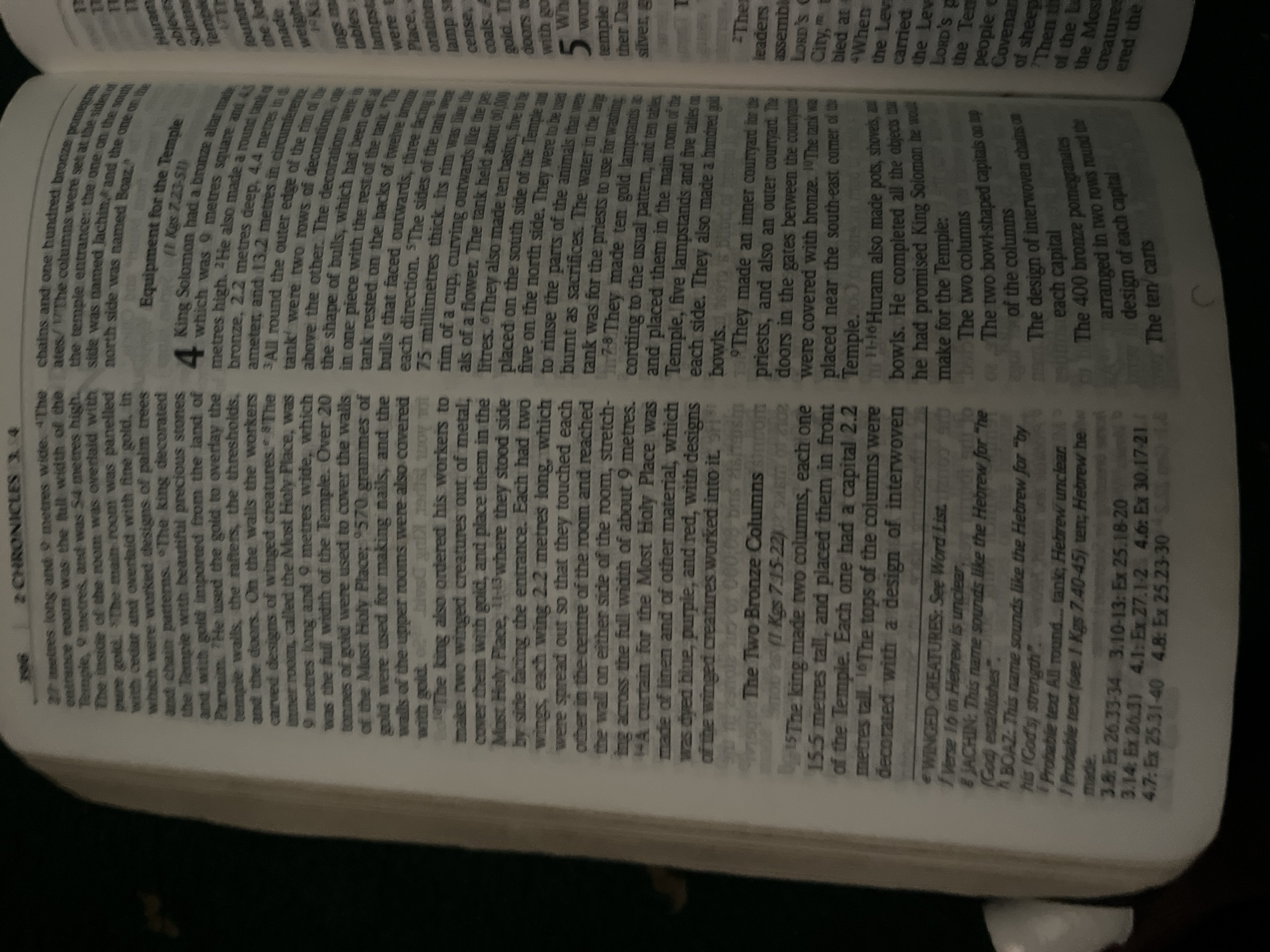 The image is a close-up, slightly angled shot of an open book, likely a Bible, with text printed in columns. The page number "396" and chapter "2 CHRONICLES" are visible at the top. The text discusses measurements, materials like gold and cedar, and religious objects. Specific verses are referenced, such as "1 Kgs 7.23-51". There are also marginal notes and a section titled "The Two Bronze Columns". The lighting suggests it was taken indoors, possibly with natural light from a window or artificial lighting. The image is focused on the text, with the surrounding environment blurred out. There is no indication of a location in Jauro Yinu, Nigeria, or any people, subjects, or specific actions beyond the depiction of religious text.