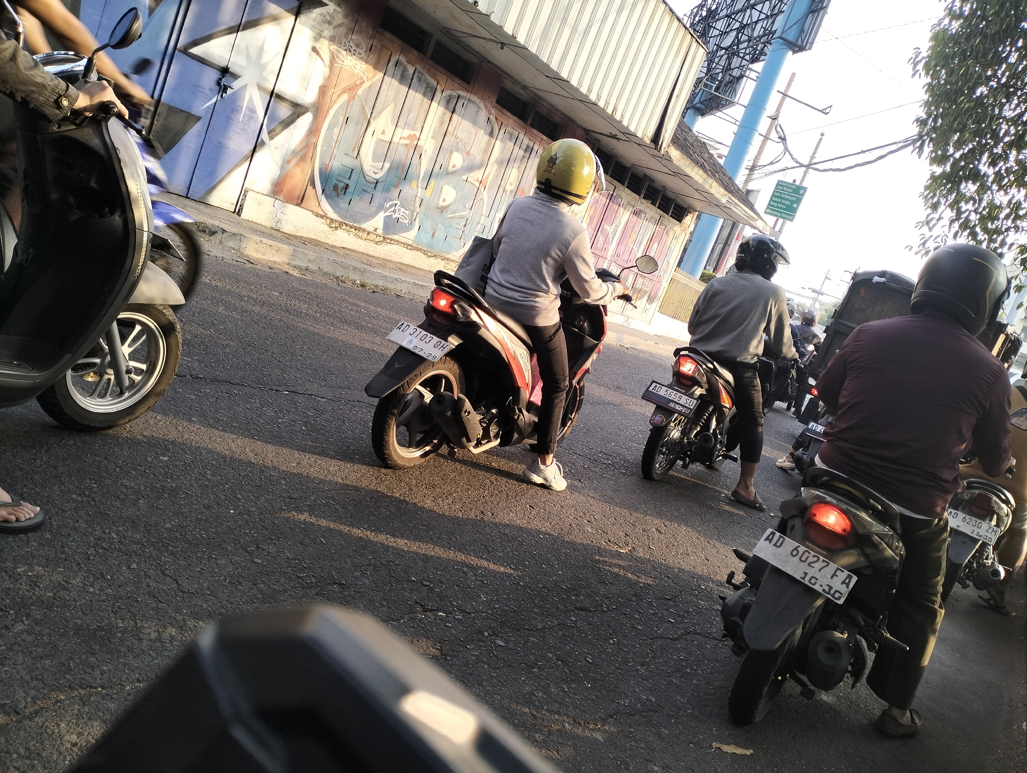The image depicts a street scene in Surakarta, Indonesia, with several motorcycles and their riders visible. In the foreground, a person wearing flip-flops is standing near a parked scooter. Further down the road, multiple motorcycles are in motion, suggesting traffic. One rider in the mid-ground is wearing a yellow helmet and a grey long-sleeved shirt, on a red and black scooter. Another rider, further back, is wearing a dark helmet and a grey shirt. A third rider in the background is wearing a dark helmet and a maroon shirt. The environment includes a building with graffiti on its facade and a roadside with trees and overhead wires. The lighting suggests it is late afternoon or early evening, with long shadows cast across the asphalt road. Several motorcycles have license plates that appear to be Indonesian. For example, one license plate reads "AD 3103 DH" and another reads "AD 6027 FA."