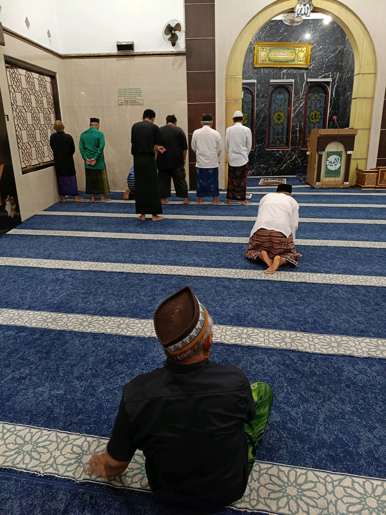 The image shows the interior of a mosque in Maguwoharjo, Indonesia. The mosque features blue carpet with white and light blue patterned stripes. Several men are present, most of whom are standing in prayer formation, facing the front of the mosque. One man is kneeling in prayer on the carpet. The architecture includes arched doorways and decorative elements on the walls. A podium with religious inscriptions is visible. The overall scene depicts a religious gathering during prayer.