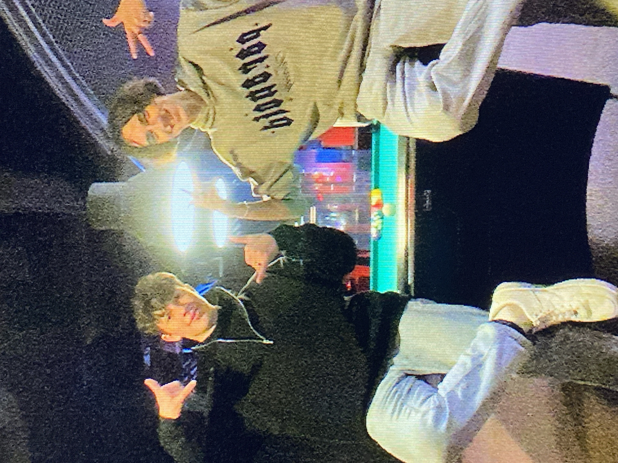 Two young men are posing for a photo in front of a pool table. The man on the left wears a black zip-up hoodie and light-colored jeans. The man on the right wears a tan hoodie with dark lettering and light-colored pants. Both men are making hand gestures. The pool table has several colored balls on it and is lit by overhead lights. The background is dark with some colorful lights visible. The image appears to be taken indoors, possibly in a bar or entertainment venue. The context suggests this may be a scene from Poznan, Poland, but this cannot be confirmed from the image alone.