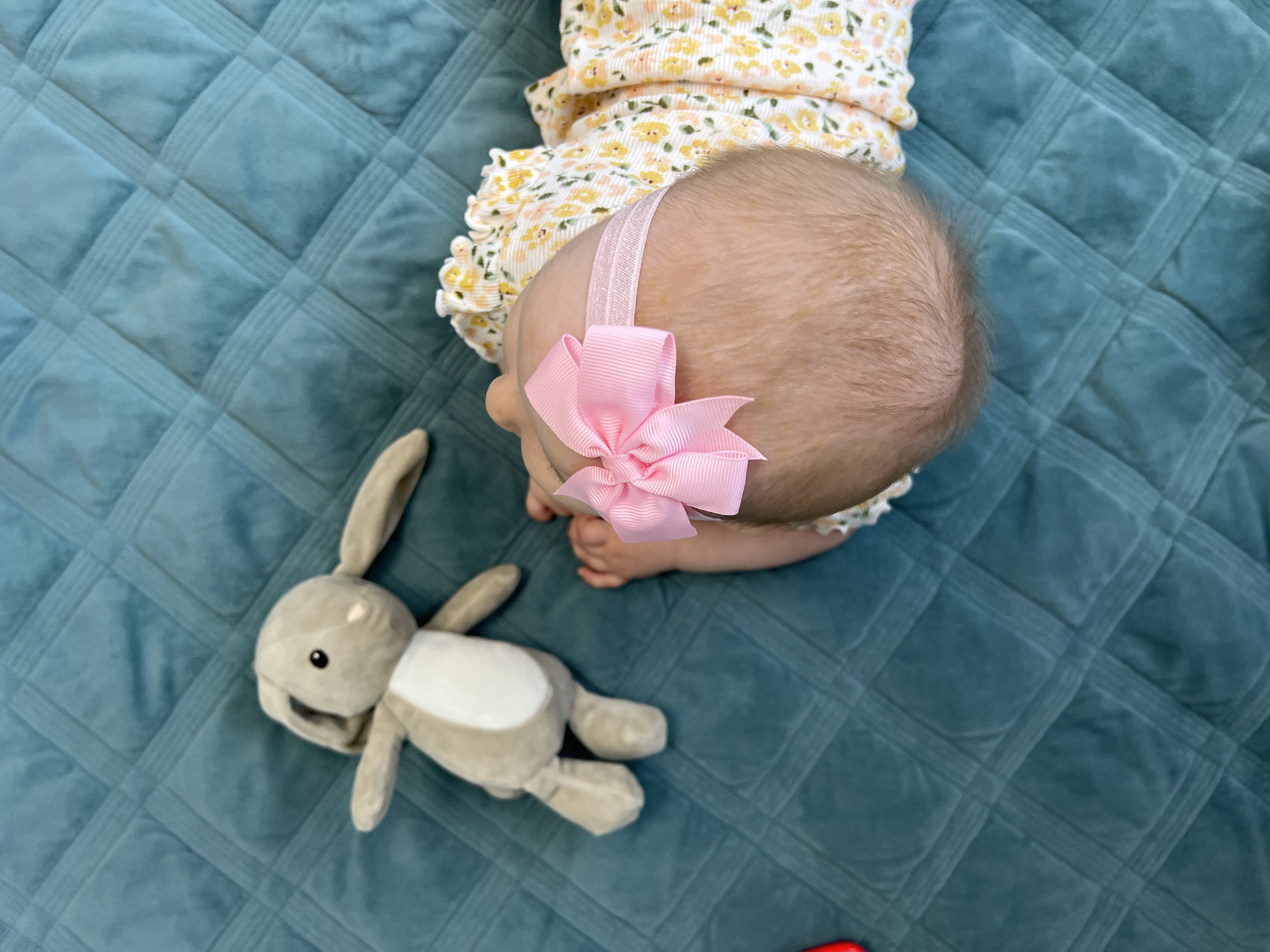 A top-down shot shows a baby on a quilted, blue-green surface next to a plush gray bunny toy. The baby wears a floral-patterned romper and a pink bow headband. The location is not discernable.