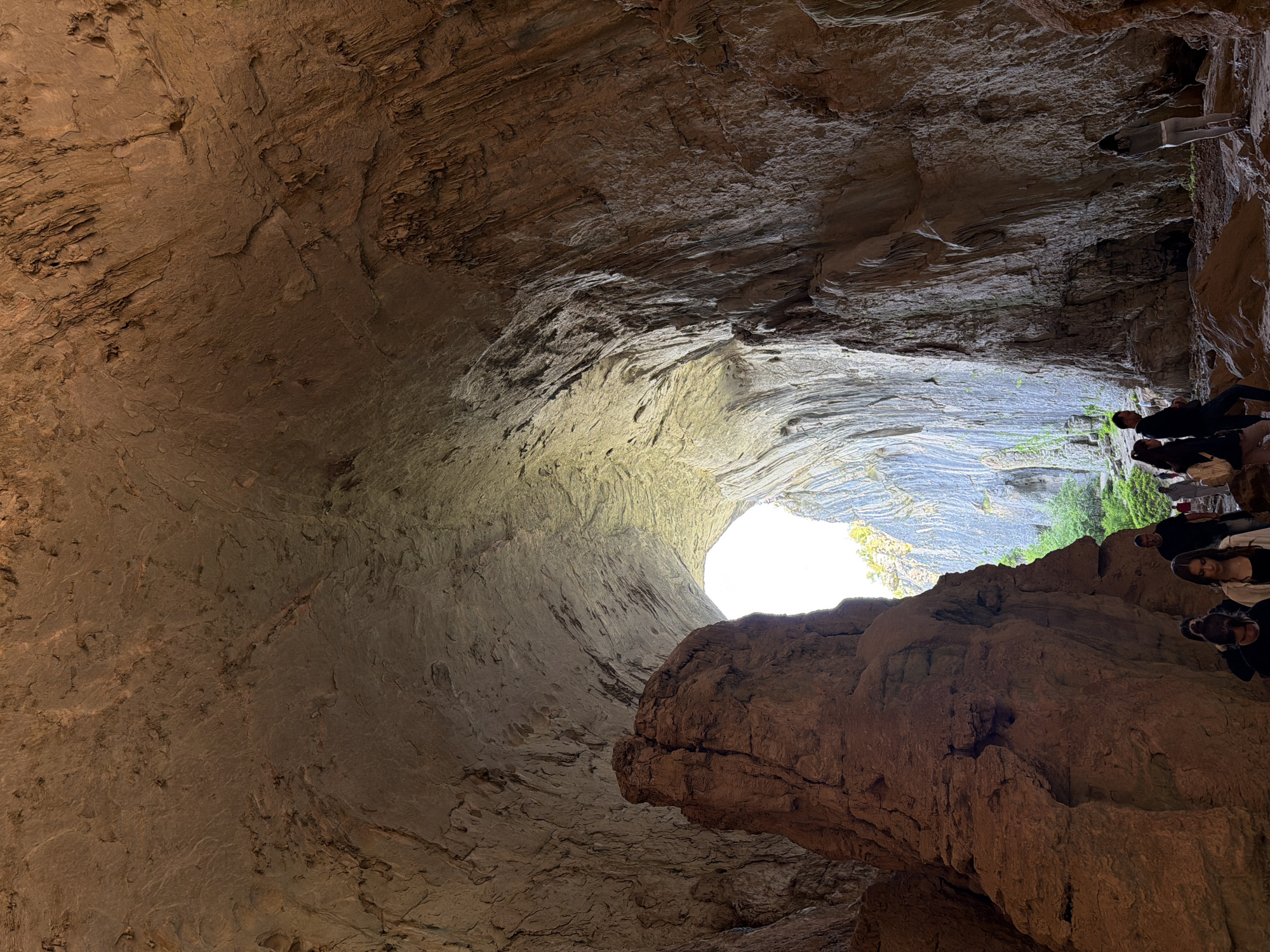 This image shows the Devetashka Cave in Karlukovo, Bulgaria. The cave has a large opening allowing daylight in. A group of people are visible near the exit of the cave. The cave walls are primarily made of light brown rock.