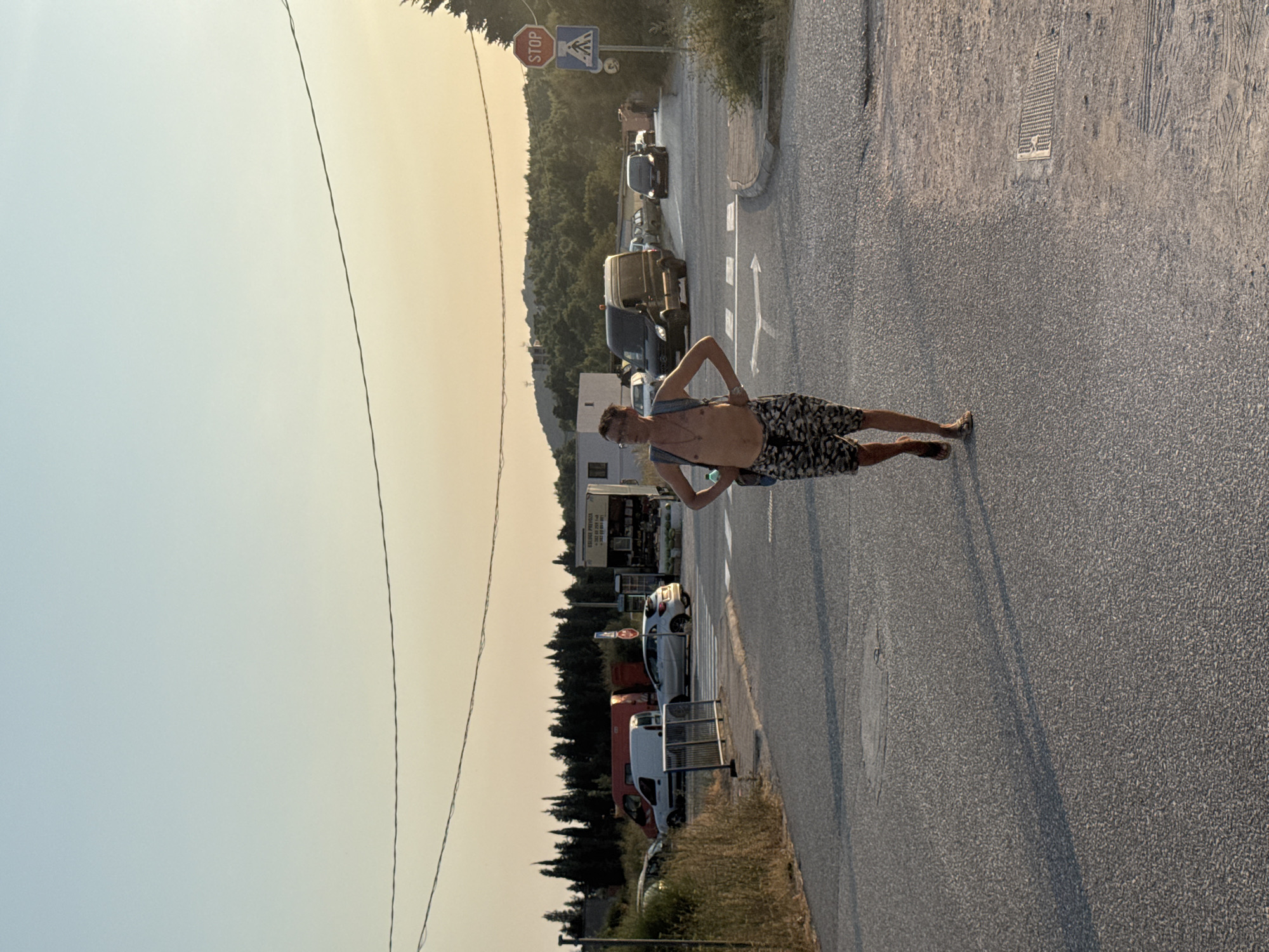The image shows a man walking across a road at a junction in Sutomore, Montenegro. The man is shirtless, wearing camouflage shorts and sandals, and carrying a backpack. He is looking towards the camera with his hands on his hips. The road is asphalt and has white directional arrows painted on it. In the background, there are several cars and vans parked or driving, and a building with a sign that reads "USLUGE PREVOZA". There are also trees and hills in the distance. The sky is clear with a warm, hazy glow, suggesting it is either early morning or late afternoon. A stop sign is visible on a pole to the right of the road.