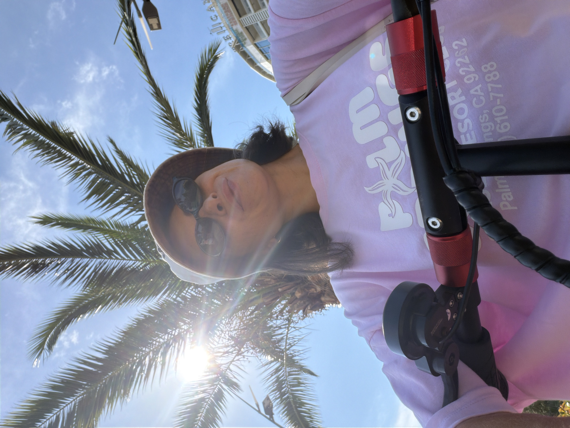 In a selfie-style shot, a person wearing a tan bucket hat and sunglasses is seen from the chest up, with the camera angled upwards. They are wearing a light pink long-sleeved shirt with a white graphic print on the front that says "Palm Resort" and a phone number and address. The person is holding onto the handlebars of an electric scooter, which are black with red accents and a black bell. A light-colored shoulder bag is visible over their right shoulder.
The background features a bright blue sky with some scattered clouds and the sun shining brightly, creating lens flare. Palm tree fronds are visible in the upper left and center of the image, suggesting a tropical or warm climate. To the right, a building with a curved facade and balconies is partially visible, with the letters "CAN" and possibly "RESORT" visible at the top, indicating a hotel or resort. There are also streetlights visible in the background.
The image does not contain any information or visual cues that place the location in Budva, Montenegro.
