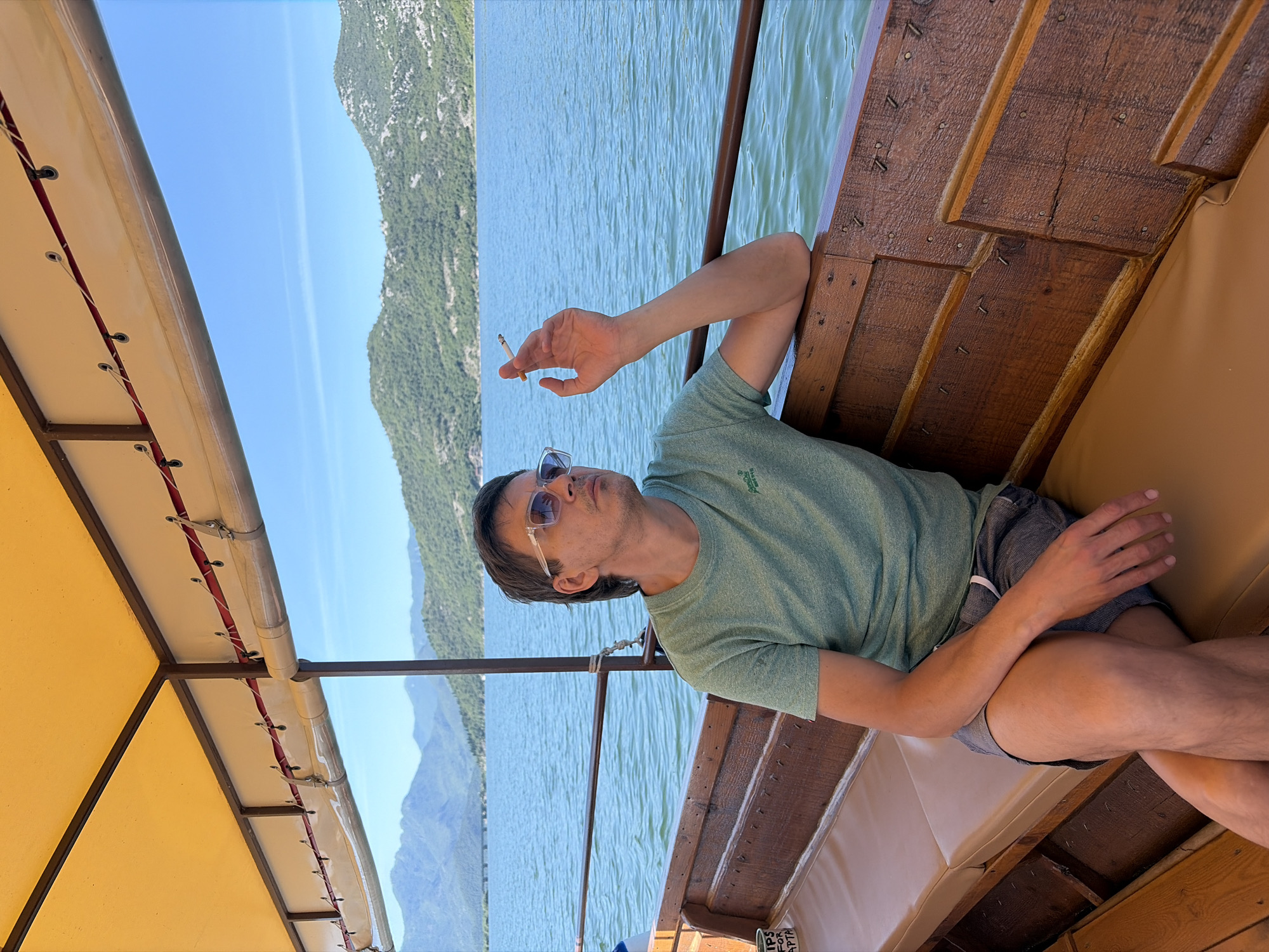 A single adult male is seated on a wooden boat, observed from the chest up on the right side of the frame. He wears a light green short-sleeved t-shirt, patterned shorts, and clear-framed sunglasses. His left hand, bent at the elbow and resting on the boat's wooden railing, holds a lit cigarette. His gaze is directed towards the left (viewer's left), across a large body of water. The boat features wooden paneling, light-colored upholstered benches, and a yellow canvas canopy supported by dark metal poles. Below the canopy, a white fabric lining with red cording and clear plastic roll-up panels is visible. To the immediate left of the man, a small container labeled "TIPS FOR CAPTA" is situated on a small shelf.
The background depicts a vast, calm body of blue-green water under a clear, bright blue sky. Across the water, a long, low bridge with multiple spans is visible in the middle distance. Beyond the water and bridge, a series of green, tree-covered mountains rise under the clear sky. The scene takes place in the vicinity of Vranjina, Montenegro.