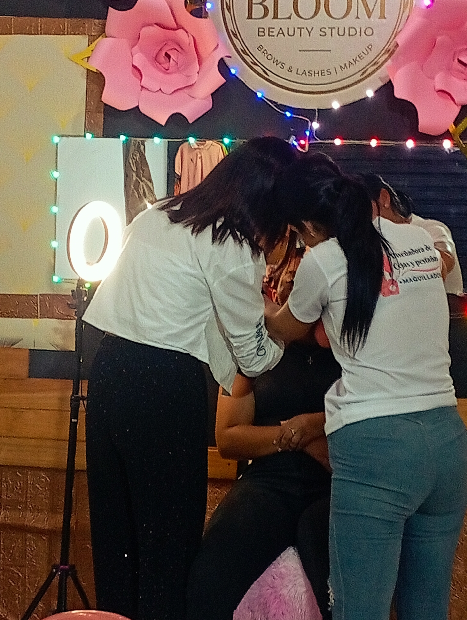 The image captures an indoor scene at "Bloom Beauty Studio" in Porlamar, Venezuela. Two women wearing white shirts and dark pants are attending to a seated person. The woman on the left is wearing black pants and has the word "BRAND" on the sleeves of her shirt. The woman on the right is wearing blue jeans and her shirt has a logo with "Delineadora de cejas y pestañas" and "MAQUILLADO" written on it. In the background, there is a lighted sign for the "Bloom Beauty Studio" and large artificial pink flowers. A ring light on a tripod stands to the left.
