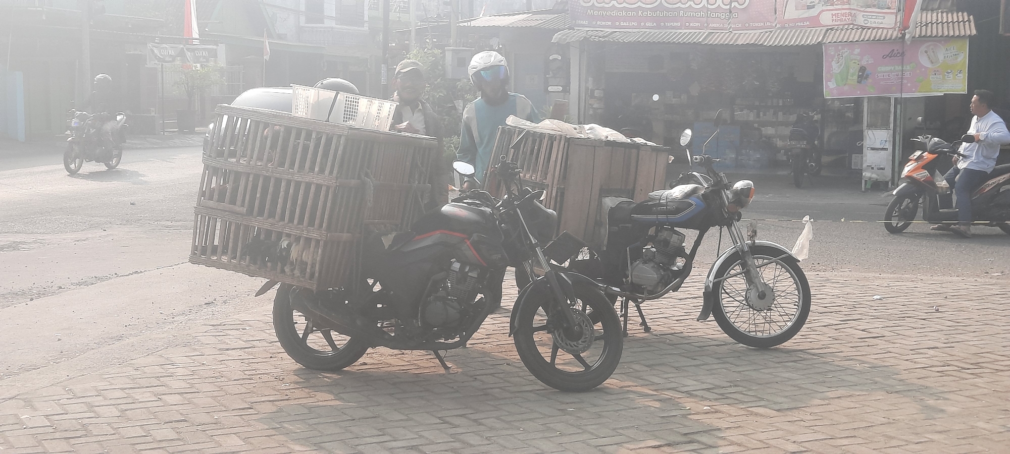 In the city of Selopuro, Indonesia, a hazy street scene unfolds with several motorcycles and their riders. In the foreground, two motorcycles are parked side-by-side. The motorcycle on the left is black and has a large wooden cage attached to its rear, filled with what appear to be poultry. A man wearing a cap and a light-colored shirt is standing next to this motorcycle, looking towards the right. To the right of this motorcycle, a second motorcycle is parked. This motorcycle is black with blue accents and also has a wooden crate on its rear. A man wearing a blue and light-colored shirt and a white helmet is seated on this motorcycle. In the background, a light orange scooter is being ridden by a man in a white shirt and blue pants. Further in the background, other motorcycles and buildings lining the street are visible, obscured by haze. The ground is paved with brickwork in the foreground.