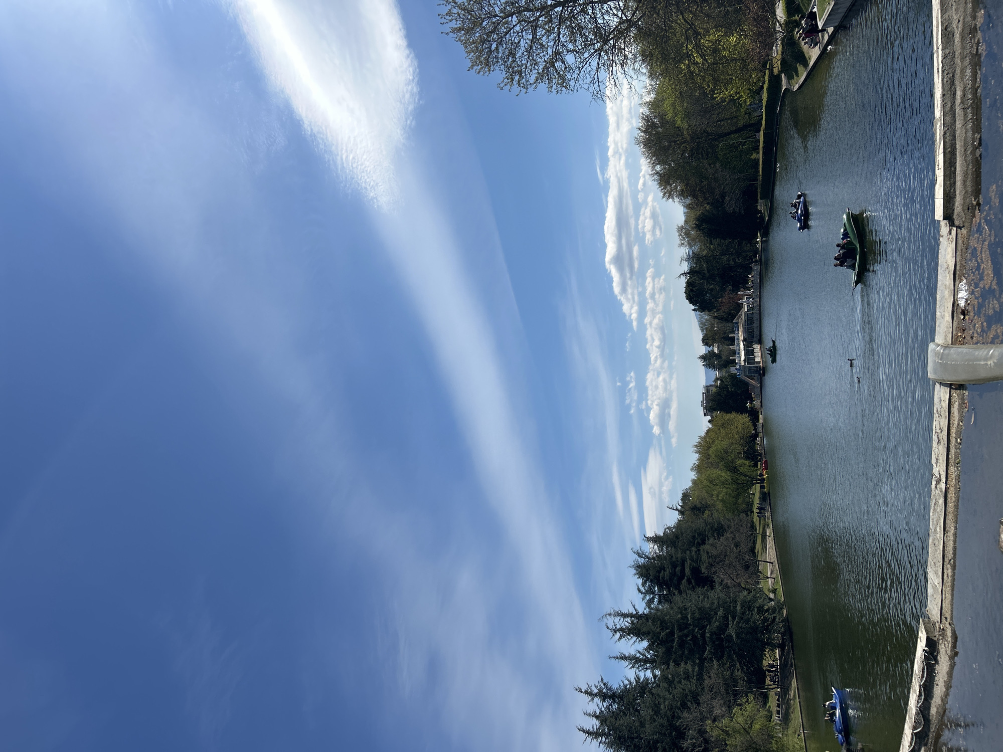 The image depicts a body of water, likely a lake or pond, with trees lining the banks. Several paddle boats with occupants are visible on the water. People are also present along the edges of the water, suggesting a park-like setting. The sky is blue with streaky clouds. The location is in the city of Unknown, Unknown.