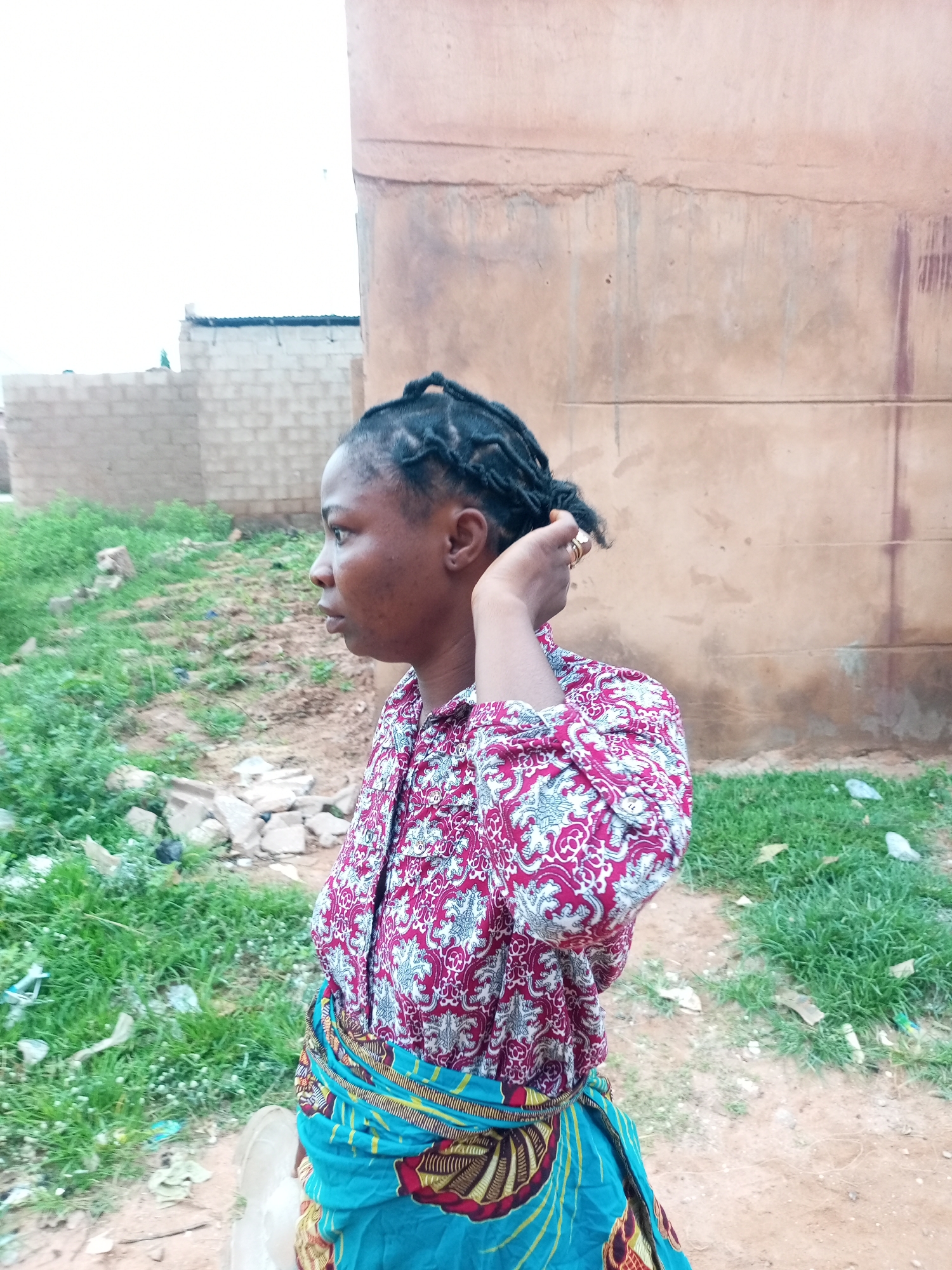 A medium shot captured outdoors features a Black woman in profile, facing left. Her dark hair is braided into small cornrows, and she is touching the back of her head with her right hand, wearing two rings on her fingers. She is dressed in a long-sleeved button-down shirt with a red and white pattern. Around her waist, she wears a blue fabric with a patterned sash. The background shows a rough, beige wall of a building. To the left of the woman, there is a patch of green grass, some scattered debris, and a brick wall. The overall environment suggests a somewhat rural or developing urban area. The image's metadata does not specify a location in Tempure, Nigeria.