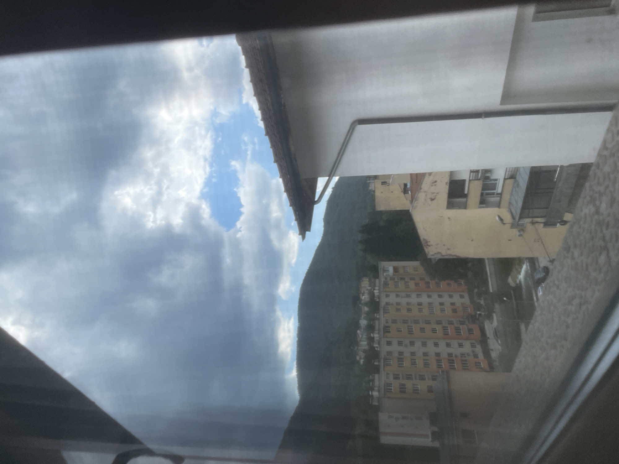 The image presents an exterior view from Vlahovo, Bulgaria, captured through a window, evident by reflections and framing on the left, top, and bottom edges. The upper portion of the scene is dominated by a cloudy sky, featuring heavy, dark gray cumulus clouds primarily on the left and center, transitioning to a patch of clear blue sky with lighter, scattered white clouds in the upper right quadrant. In the background, a dense, forested mountain range extends across the horizon.
The mid-ground features a cluster of residential buildings nestled at the base of the mountains. A prominent multi-story apartment block, characterized by a light orange and beige facade with numerous windows, is centrally located on the left. In the immediate right foreground, a section of a white building with a red terracotta tile roof and an exposed downspout system is visible, partially obscuring the distant landscape. Adjacent to and below this structure are parts of other yellow and beige buildings, some displaying balconies. A partial view of a street or parking area with at least one dark colored vehicle is discernible beneath the central apartment block. No people, specific actions, or interactions are observable within the frame.