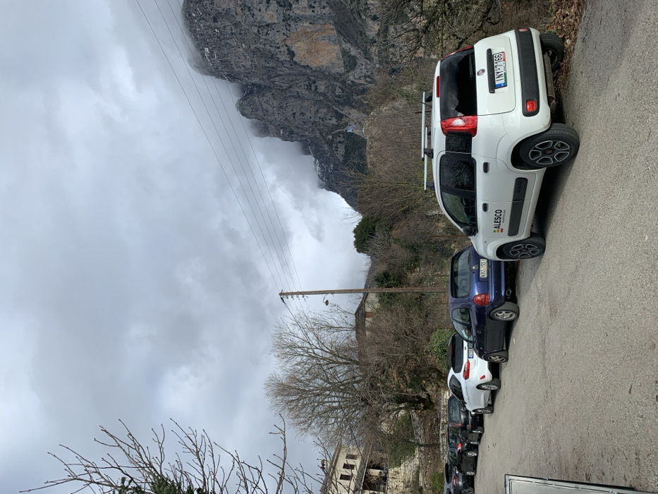 The image, taken in Vikos, Greece, is oriented vertically and depicts a mountainous landscape with a line of parked cars along a paved road. The sky is heavily overcast with thick grey clouds, and mist or low clouds partially obscure the upper parts of the rocky mountains in the background.
In the foreground, a row of cars is parked facing to the left (if the image were rotated correctly). The most prominent vehicle is a white Fiat Panda with roof rails, visible in the upper right. Its rear license plate reads "INY 1166" and on its side, a logo with the text "ALESCO" is visible. Immediately behind it is a dark blue car, possibly an Opel Corsa, with a license plate reading "VBZ 055". Several other cars, mostly dark-colored, are parked further down the line, partially obscured.
The road runs along the side of a steep incline, which is covered with bare deciduous trees, suggesting late winter or early spring. Some green conifers are also present. A wooden utility pole with power lines runs parallel to the road. In the lower left quadrant of the image, parts of a traditional stone building with a light-colored roof are visible through the branches of bare trees. The overall atmosphere is quiet and somewhat muted due to the cloudy weather.