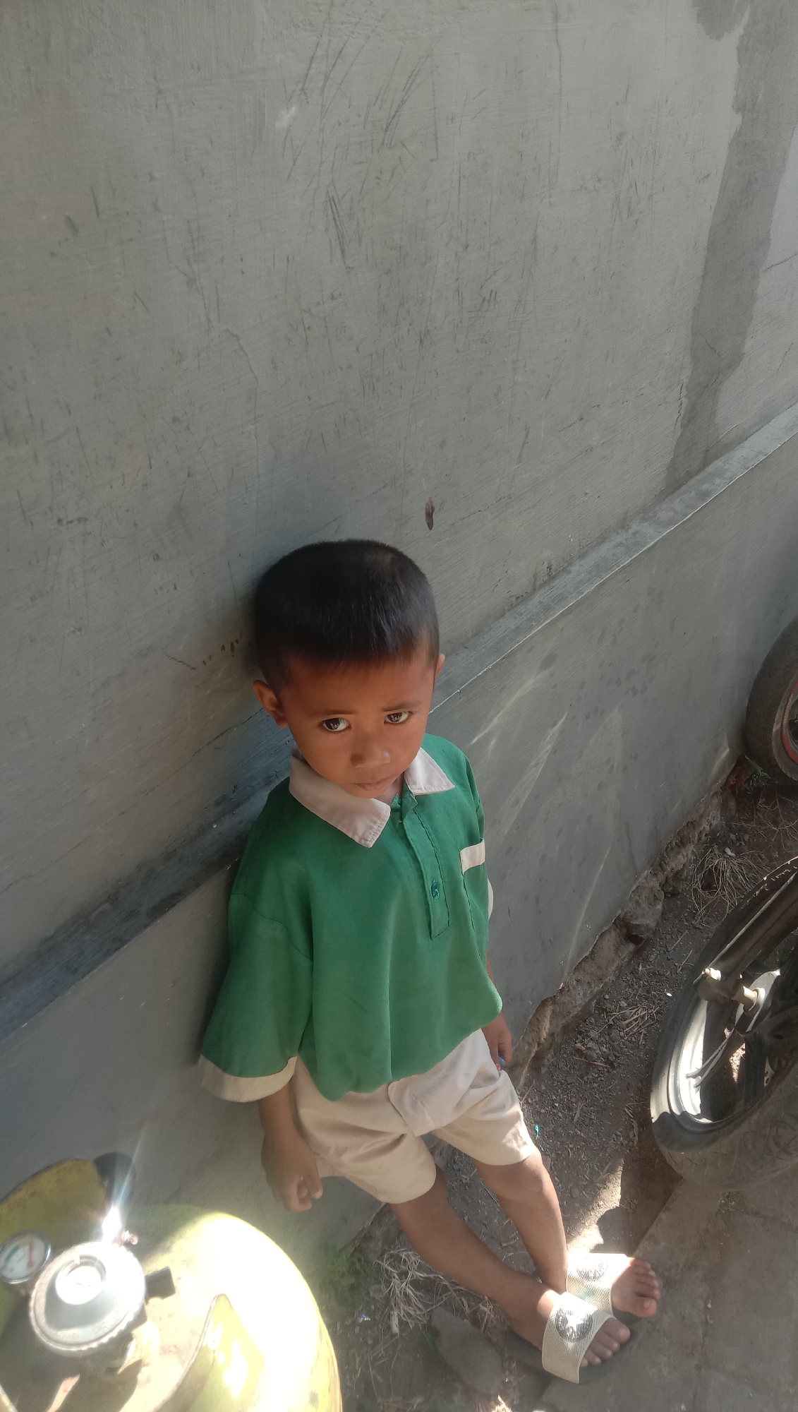 A young boy with dark hair and brown eyes is standing with his back against a gray concrete wall. He is wearing a green polo shirt with a white collar and a white stripe on the left chest, and light tan shorts. His feet are bare except for white sandals with a black and white patterned strap. To the left of the boy, a portion of a yellow gas tank is visible with a pressure gauge. To the right, a black motorcycle wheel and part of its frame are visible. The ground around the boy is dirt and dry grass. The scene is outdoors, likely in a residential area of Karanganyar, Indonesia, given the presence of common household items. The lighting suggests it is daytime, with shadows cast on the wall and ground.