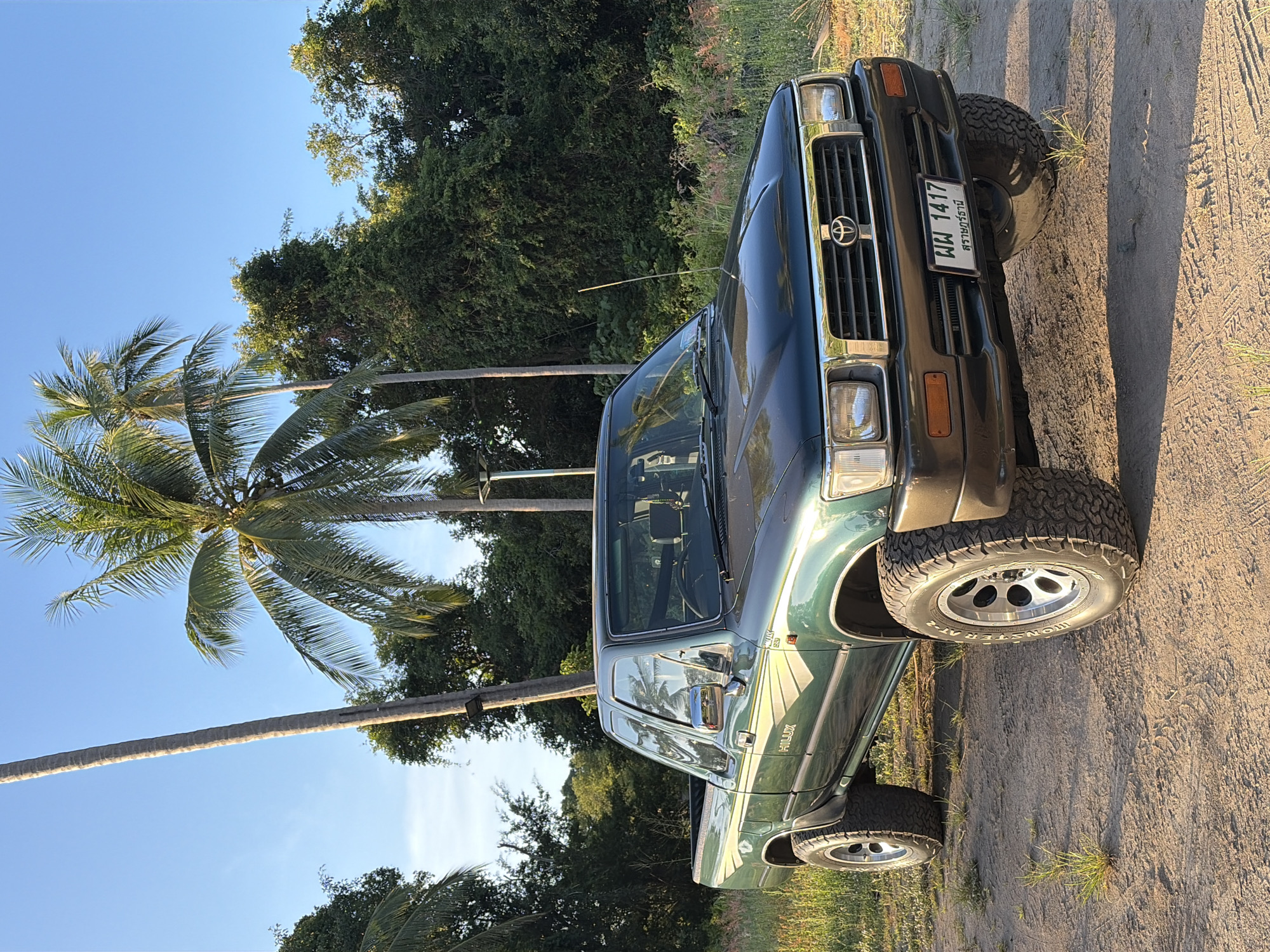 This media file is a photograph of a dark green Toyota Hilux pickup truck parked on a dirt road. The truck is oriented vertically in the frame. It has a chrome grill with the Toyota logo, square headlights, and a visible license plate. The license plate reads "ผผ 1417" and has Thai text below it. The truck appears to have off-road tires.
The setting is outdoors, with lush green trees and palm trees visible in the background. The sky is a clear, bright blue, suggesting it is daytime and the weather is fair. The sun is casting shadows, indicating it is likely late afternoon or early morning. The ground is dry and dusty, typical of a rural or unpaved road.
There are no people visible in the image. The focus is solely on the truck and its surroundings. The overall mood is peaceful and serene, capturing a moment in a tropical or subtropical environment. The location context is Baan Taling Ngam, Thailand, which aligns with the presence of palm trees and the style of the license plate.