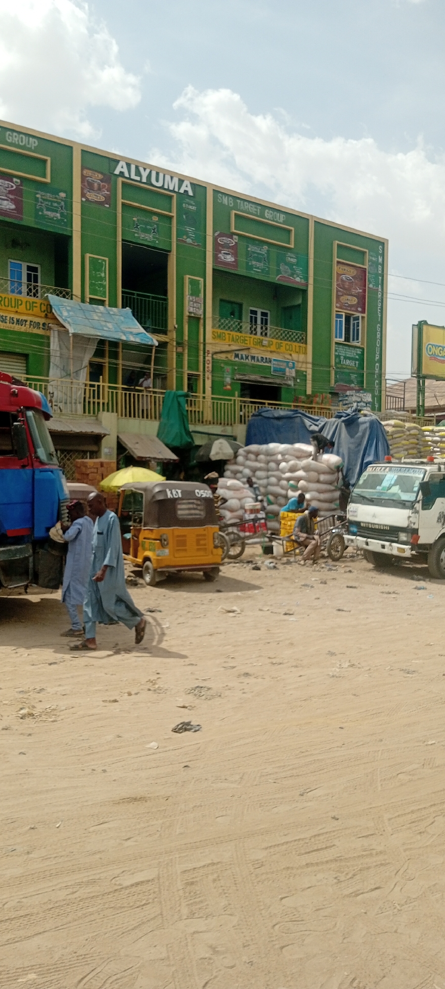 The image shows a street scene in Sabon Gari West, Nigeria, with a large green building labeled "ALYUMA SMB TARGET GROUP" dominating the background. In front of the building, there is a bustling scene with various activities. Two men wearing blue robes walk along a sandy street. There is an auto rickshaw with the license plate "KBT 0500," parked beside them. A stack of white bags covered by a tarp sits near a parked white Mitsubishi truck. In the background, a person is sitting on a handcart. Other signs visible include "MAKWARAN" and "ONGA".