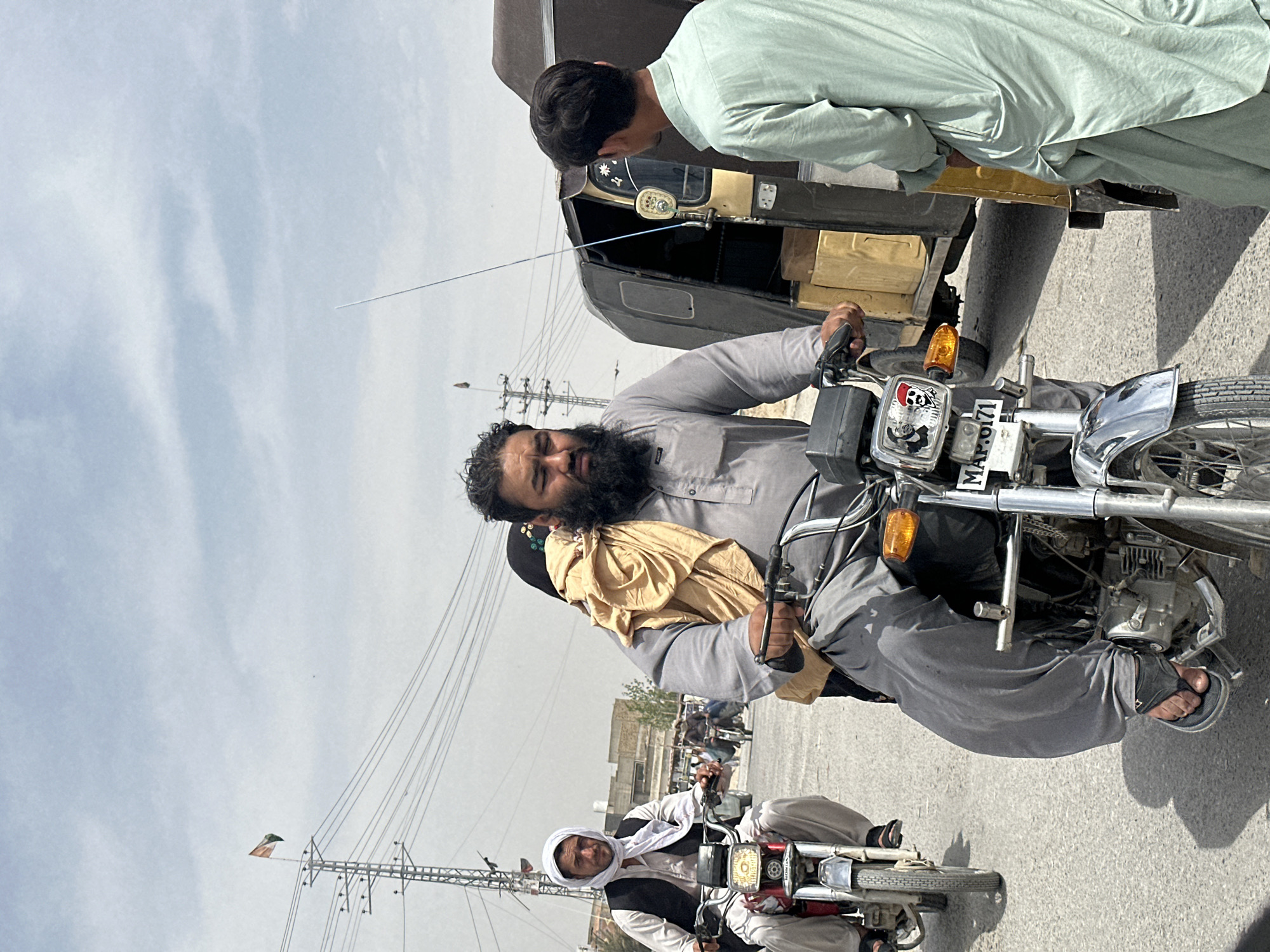 A man on a motorcycle is looking backward and talking to someone standing by an auto rickshaw in a city street in Unknown, Unknown. The man on the motorcycle is wearing a light gray shalwar kameez and sandals. He has a beard and appears to be holding something wrapped in a yellow cloth. Another motorcycle is behind him, driven by a man wearing a white head covering. Power lines are visible overhead. The sky is overcast.