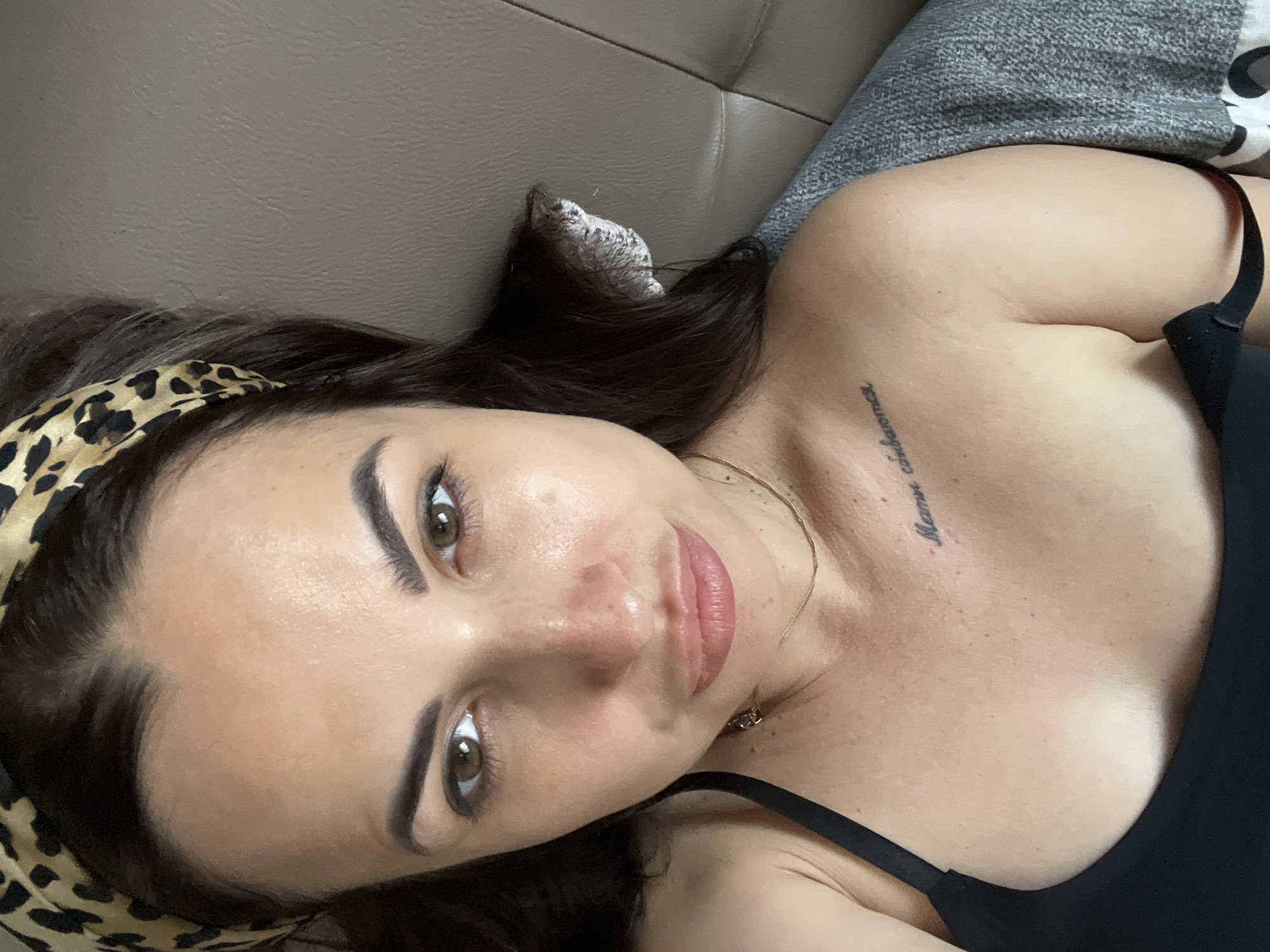 A woman with dark hair and brown eyes, wearing a black tank top and a leopard print headband, lies on a couch. She has a tattoo on her chest that reads "Мамы становятся". The image appears to be a selfie taken from a close-up perspective. The lighting is natural and soft, and the background is a textured, neutral-colored couch.