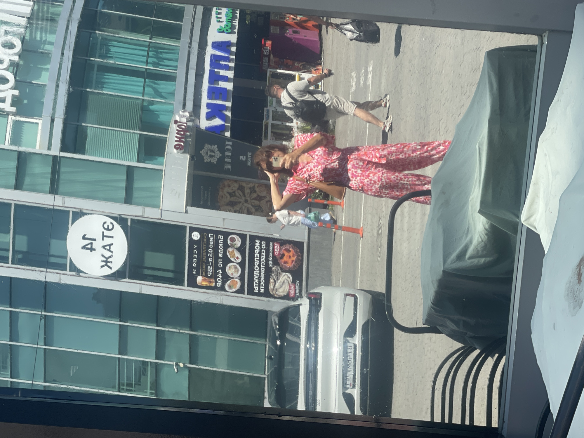 The image captures a reflection in a window, likely from a cafe or shop, showing a street scene in Sochi, Russia. In the foreground of the reflection, a woman wearing a pink floral jumpsuit and sunglasses is taking a selfie with her phone. She is standing on a paved area, with a black metal railing and a tarp-covered object to her left.
Behind her, the reflection reveals a modern building with a facade of glass windows and vertical elements. A circular sign with Cyrillic lettering, "АГ ЖАТЕ," is visible on the building. Further back, a white car is parked, with a license plate partially obscured. Another sign with Cyrillic text and images of food is mounted on the building.
Several other individuals are visible in the background of the reflection, including a man with a backpack walking away, and other pedestrians. Orange traffic cones are present on the ground. The environment appears to be an urban setting with a bright, sunny day. The reflections are slightly distorted due to the surface of the window.