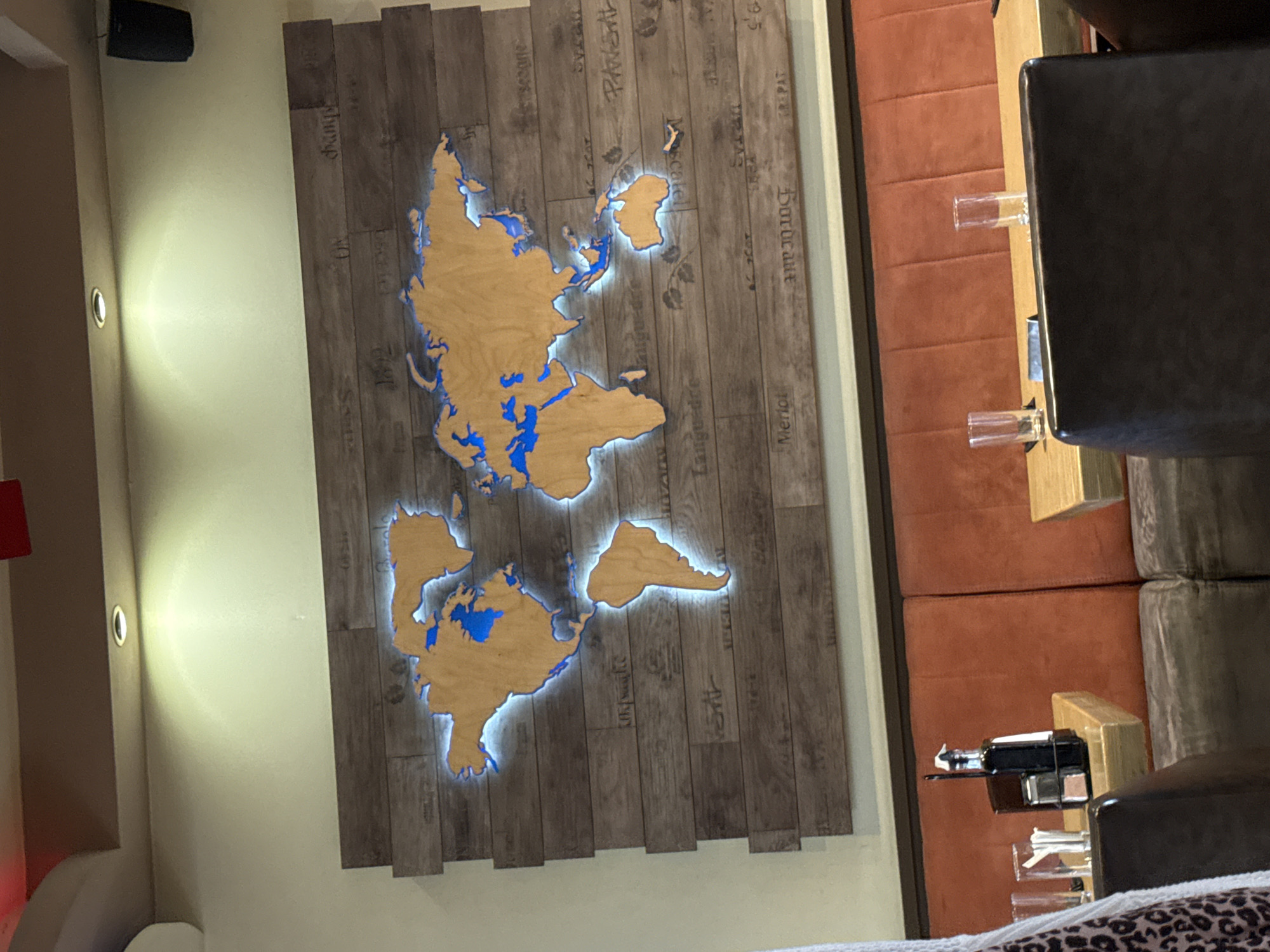 The image displays the interior of a commercial establishment, likely a restaurant or bar, in an unknown city and country. The focal point is a large, backlit wooden world map mounted on a wall covered with horizontal wooden planks of varying brown tones. The continents of the map are a light brown wood, while the oceans are a solid blue. A light blue glow emanates from behind the wooden continents, highlighting their shapes against the plank background. Various wine-related terms, including "Champagne," "Bordeaux," "Merlot," "Syrah," "Languedoc," and "Chianti," are lightly etched or printed on the wooden planks, some upright and some inverted.
Above the map, three circular recessed lights are embedded in a brown ceiling soffit, casting warm yellowish light onto the wall. A black speaker is mounted on the upper right wall. Below the map, a long upholstered booth extends across the width of the frame. The back cushions of the booth are a rust-orange color with vertical segmentation, and the seat cushions are a dark, textured gray-brown. In front of the booth, several light-colored wooden tables are visible. On the table to the left, a black caddy holds a dark bottle, a metal shaker, and a container of white stick-like items (possibly napkins or straws). Two clear drinking glasses are present on the rightmost visible table. Dark brown upholstered chairs are partially visible in the foreground, facing the tables, with a leopard-print fabric draped over the back of the leftmost chair. No people or specific actions are depicted in the scene.
