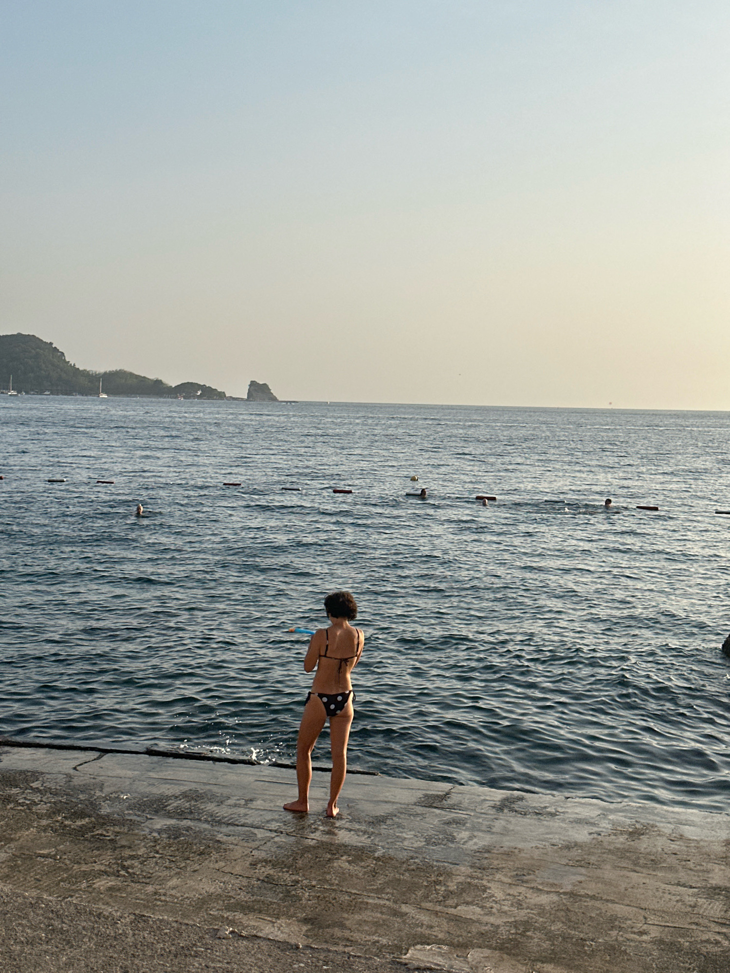 A person stands on a concrete pier facing the sea, wearing a black and white polka dot bikini. They are holding a blue object. In the distance, several people are swimming in the sea, with a line of buoys marking a swimming area. Mountains covered in greenery form the backdrop on the horizon, with two sailboats visible near the shore. The sky is a clear, pale blue, transitioning to a lighter hue near the horizon. The image appears to be taken in Budva, Montenegro, given the coastal scenery.