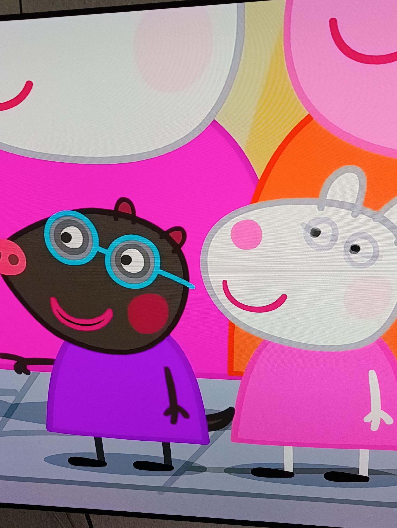 The image shows a scene from the children's cartoon "Peppa Pig." In the foreground, there is a mole character with dark brown skin, wearing blue glasses and a purple shirt. Next to the mole is a white rabbit character wearing a pink dress. In the background, there are blurred figures of other characters, possibly pigs. The setting appears to be a sidewalk or paved area, suggested by the gray lines on the ground.