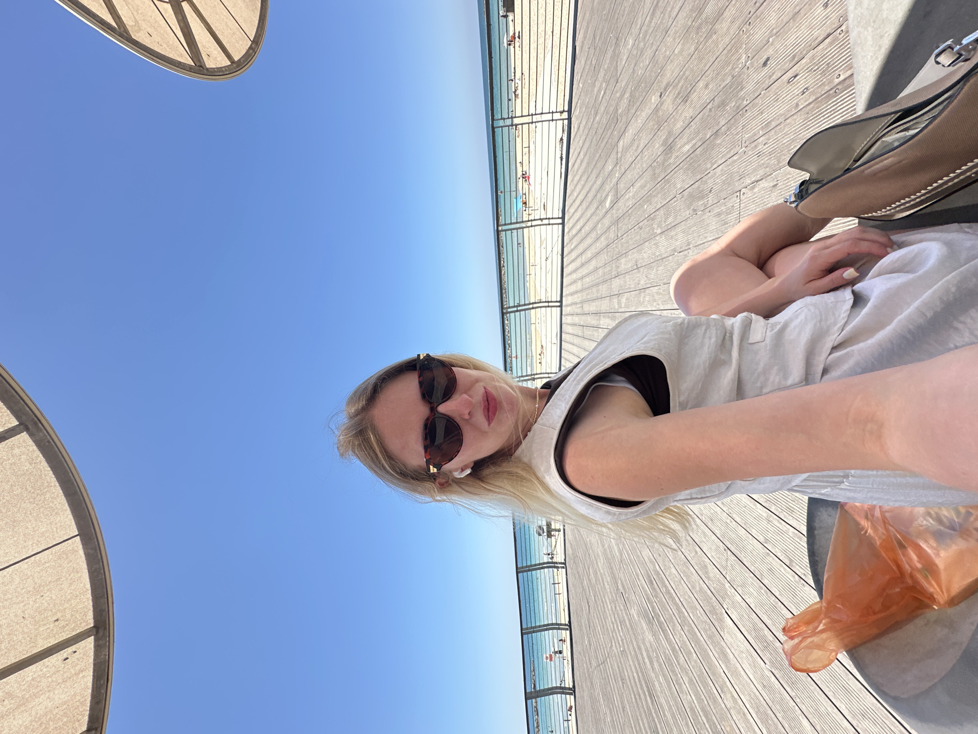 The image is a selfie taken outdoors on a bright, sunny day, depicting a coastal scene in Tel-Aviv, Israel. A clear, expansive blue sky dominates the left side of the frame, indicating excellent weather. To the right, a light-colored wooden boardwalk stretches into the distance. Between the sky and the boardwalk, a transparent railing overlooks a sandy beach with tiny figures of people enjoying the sun, and the calm blue sea beyond.
The primary subject is a young woman with long blonde hair, wearing dark sunglasses and white wireless earbuds. She has a neutral expression, looking directly at the camera, and is dressed in a light-colored, possibly linen, sleeveless top or dress. Her posture suggests she is seated or reclining, with her legs appearing crossed.
Beside her, on the wooden deck, a beige handbag is partially visible, along with a crumpled orange plastic bag. In the upper left corner, part of a large, light-colored canopy or umbrella structure provides shade. The strong sunlight and lack of clouds suggest it is midday or early afternoon. No visible text is present in the image. The overall impression is one of leisure and enjoyment in a warm, picturesque outdoor setting.