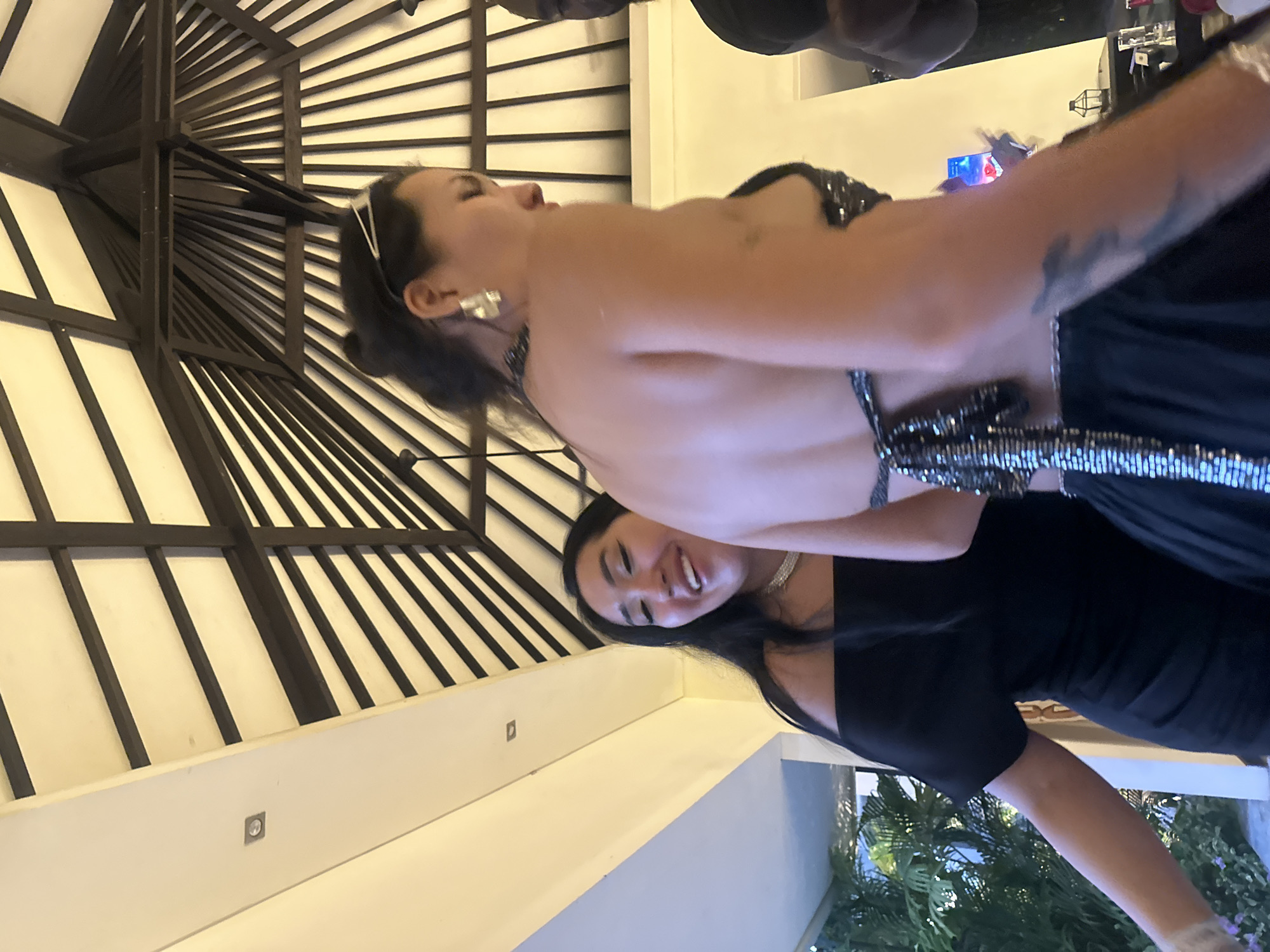 Two women stand beneath a distinctive architectural structure in what appears to be a courtyard or outdoor event space in North Kuta, Indonesia. The structure is a patterned ceiling with dark beams against a light background. The first woman wears a backless black dress with a beaded or sequined strap. The second woman wears a short-sleeved black dress with a shallow neckline. The surrounding environment includes glimpses of greenery suggesting an outdoor setting, potentially a garden or tropical landscape.