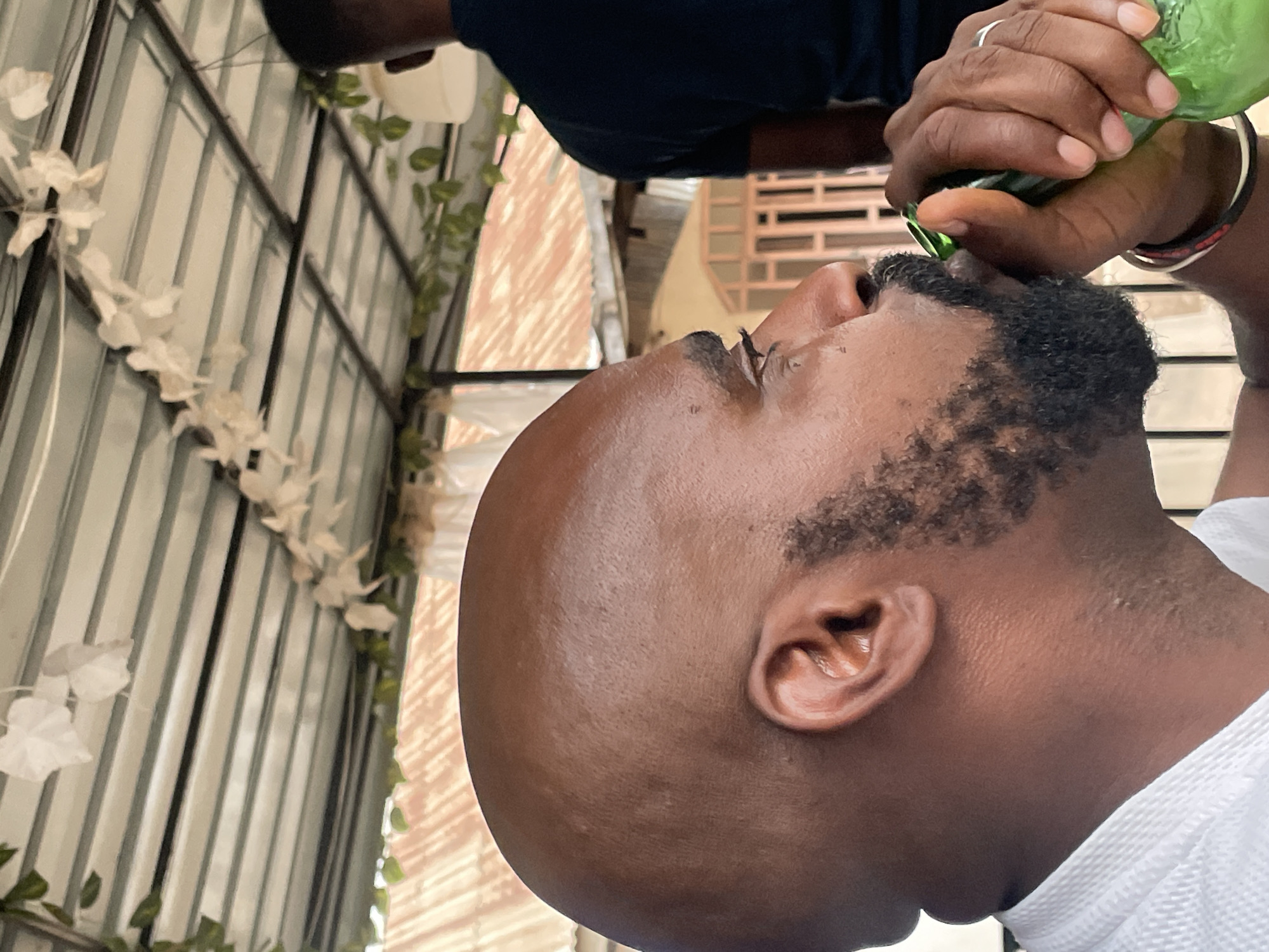 A close-up, side-profile shot depicts a dark-skinned man drinking from a green glass bottle. He has a shaved head, a beard, and is wearing a white shirt. The bottle is held to his lips by a dark-skinned hand wearing a ring and several bracelets. In the background, a wall with decorative white flowers and green vines is visible. Jos, Nigeria is not discernible from the image.