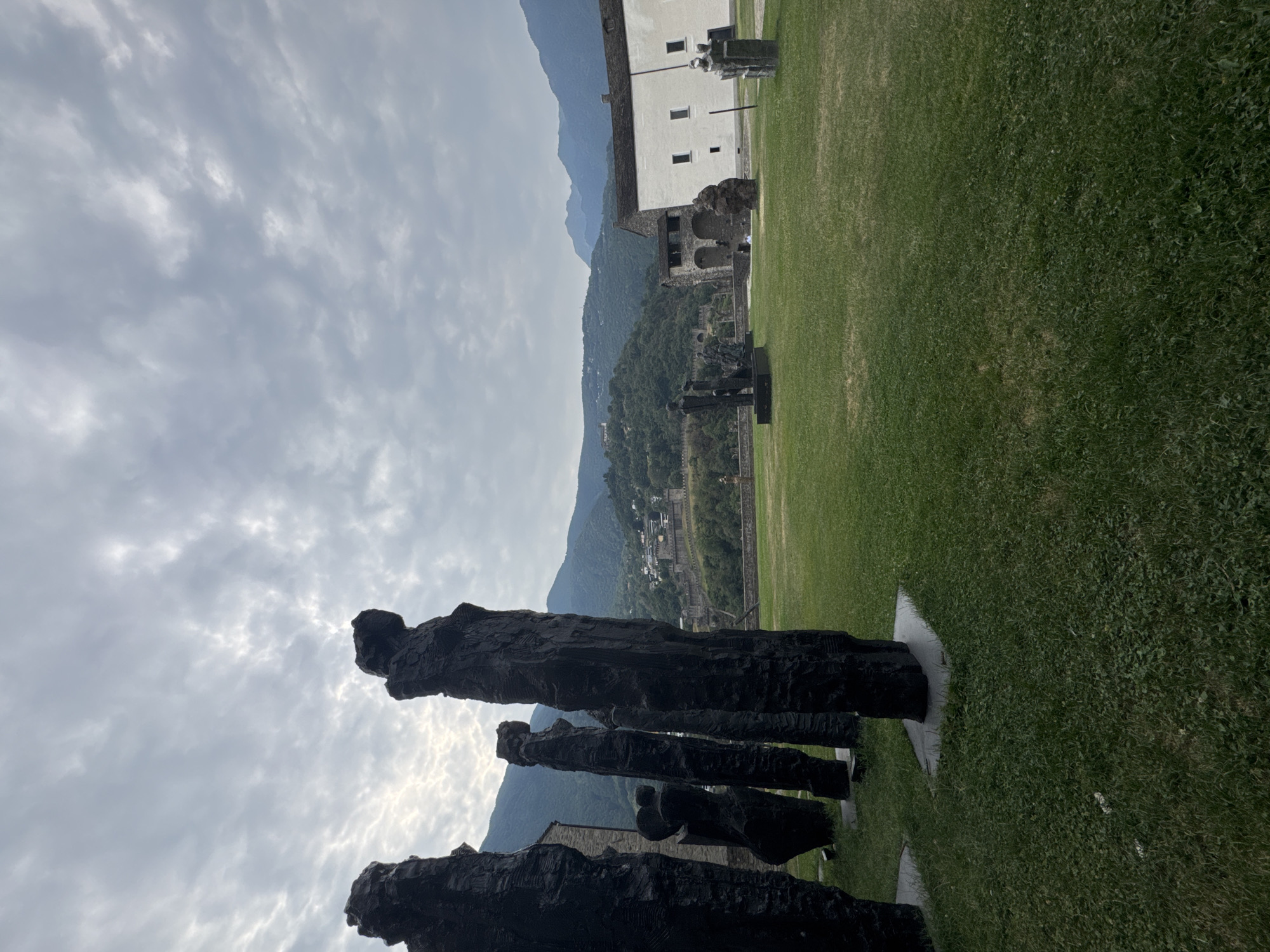 An outdoor sculpture exhibition is set against a backdrop of lush green hills and a cloudy sky. In the foreground, several dark, abstract, human-shaped sculptures stand tall on a grassy lawn. These sculptures are rough and textured, with a distinct, raw appearance. Further into the scene, the landscape opens up to reveal more sculptures scattered across the green expanse.
Behind the sculptures, a historic stone structure, likely part of a castle or fortress, is visible, nestled on a hillside. The architecture suggests an older construction, with arches and battlements. Adjacent to this historic building is a more modern, white building with a dark roof and multiple windows.
The surrounding environment is characterized by rolling hills covered in dense green vegetation, suggesting a mountainous region. The sky above is overcast with scattered clouds, indicating a potentially temperate climate. The overall impression is one of an art installation integrated into a historical and natural landscape, possibly within the grounds of Castel Grande in Bellinzona, Switzerland.